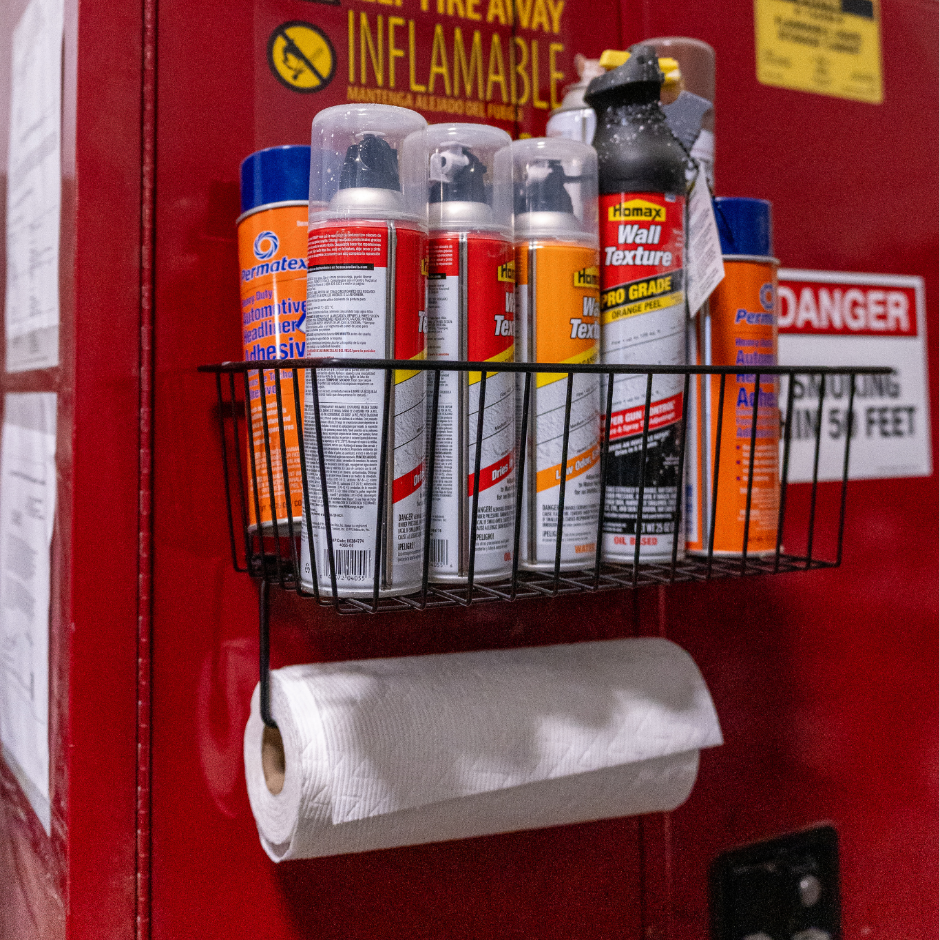 Magnetic basket mounted on a red steel storage cabinet in a workshop, holding spray adhesive and wall texture cans, with a hook below storing a white paper towel roll for organized storage and easy access