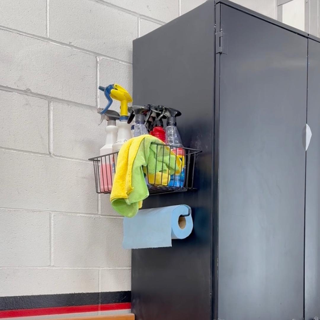 Magnetic basket mounted on a black steel cabinet in a workshop, used for storage and organization of spray bottles, cleaning supplies, and microfiber towels, with a hook below holding a blue paper towel roll for easy access.