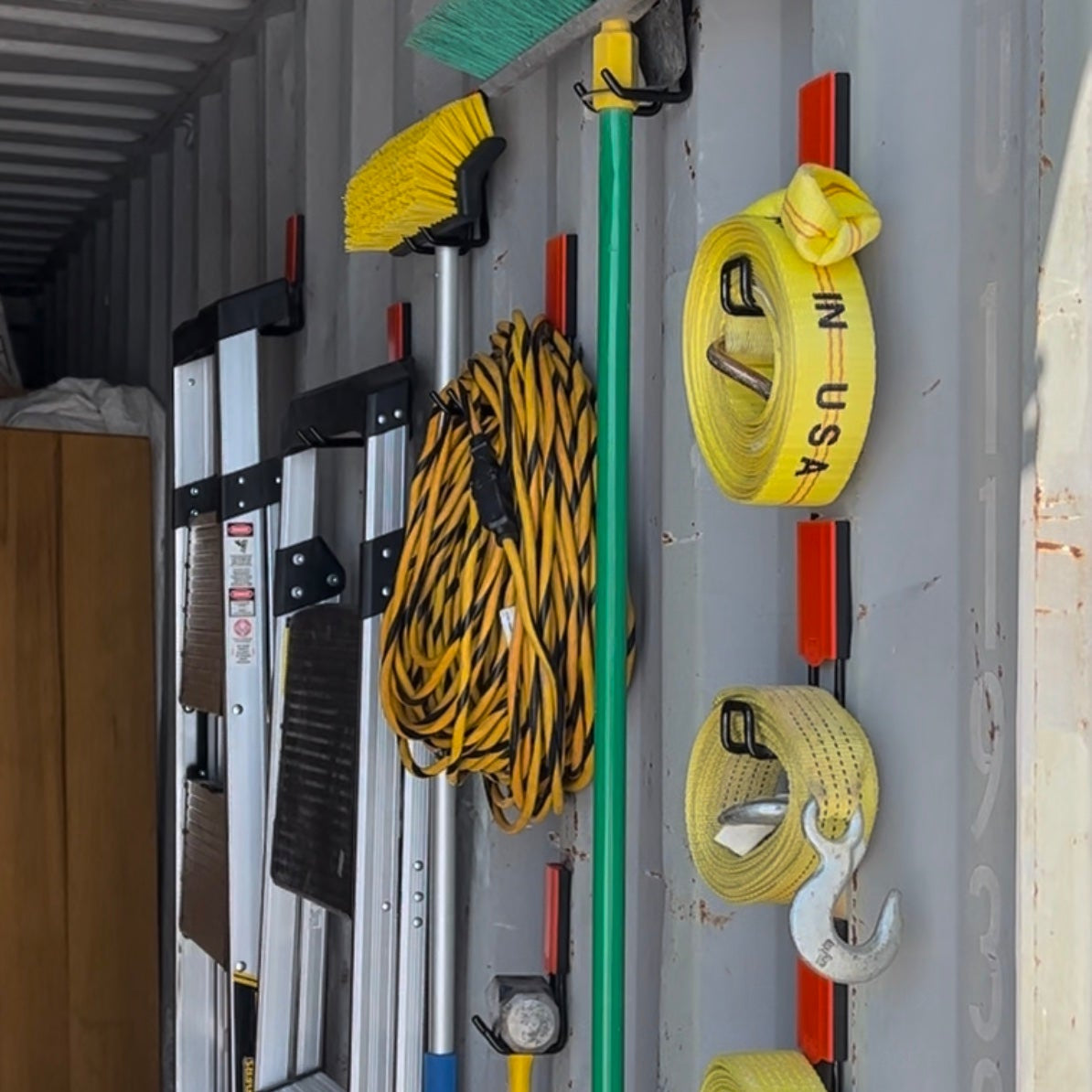 Heavy-duty magnetic hook holding brooms, extension cords, ladders, and tow straps on a storage container wall for organized tool storage and easy access.