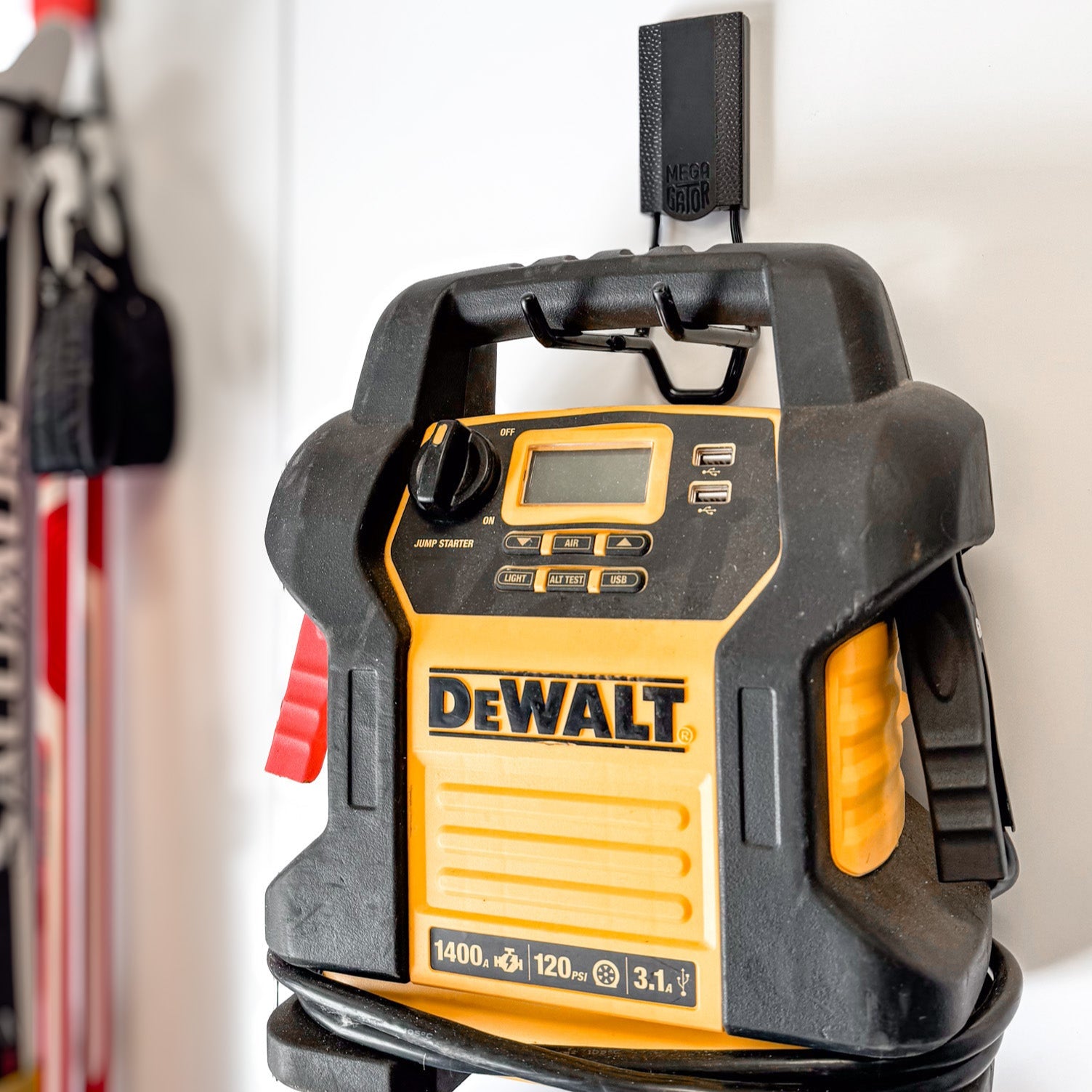 A DEWALT jump starter is hanging by its handle on a black Gator Magnetics magnetic hook attached to a white painted wall, demonstrating a wall item holder for easy and secure storage in a workshop or garage.