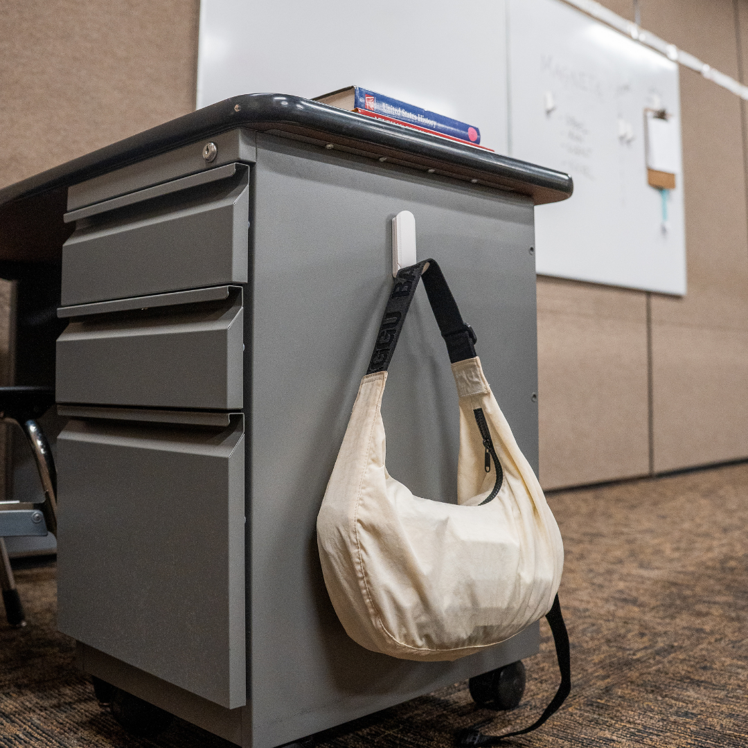 White magnetic hook attached to a gray metal desk holding a cream-colored shoulder bag, providing convenient classroom or office storage.