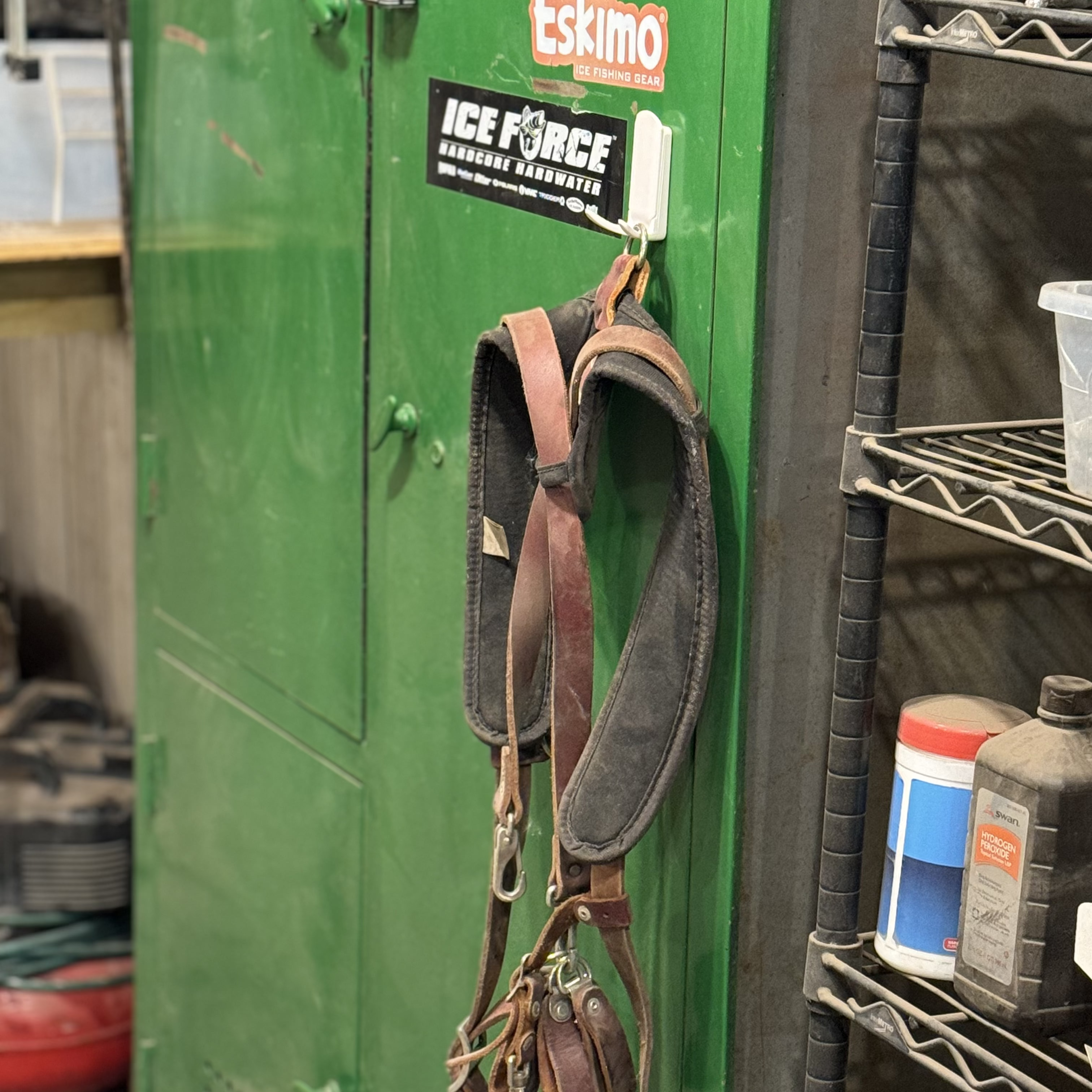 A brown leather harness is hanging from a white Gator Magnetics magnetic hook attached to a green steel cabinet door in a workshop setting, with a black metal wire shelving unit holding containers nearby and a faint glimpse of a white small basket in the background.