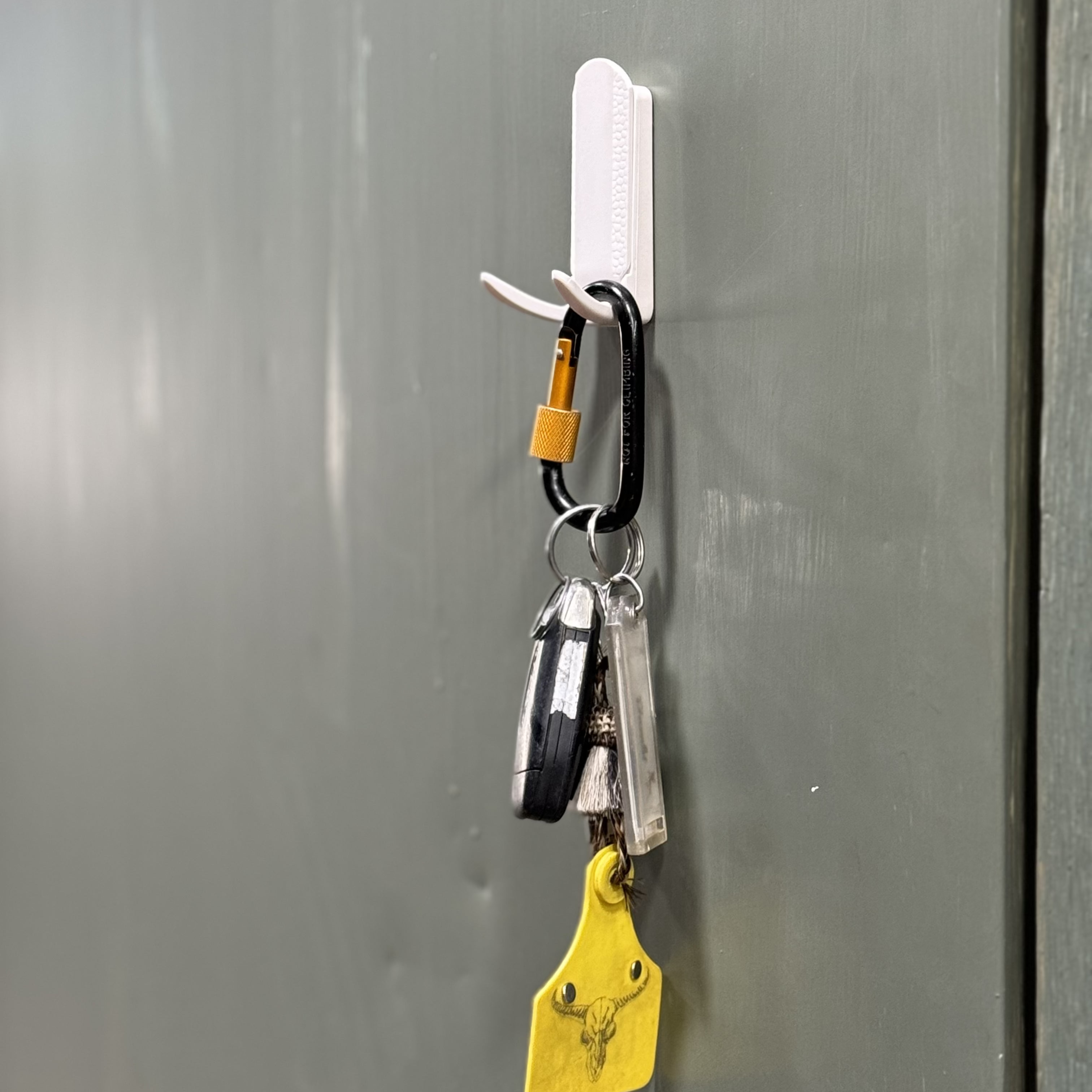 White magnetic hook on a gray steel surface holding a set of keys with a carabiner, key fob, and yellow key tag, providing easy and accessible key storage.