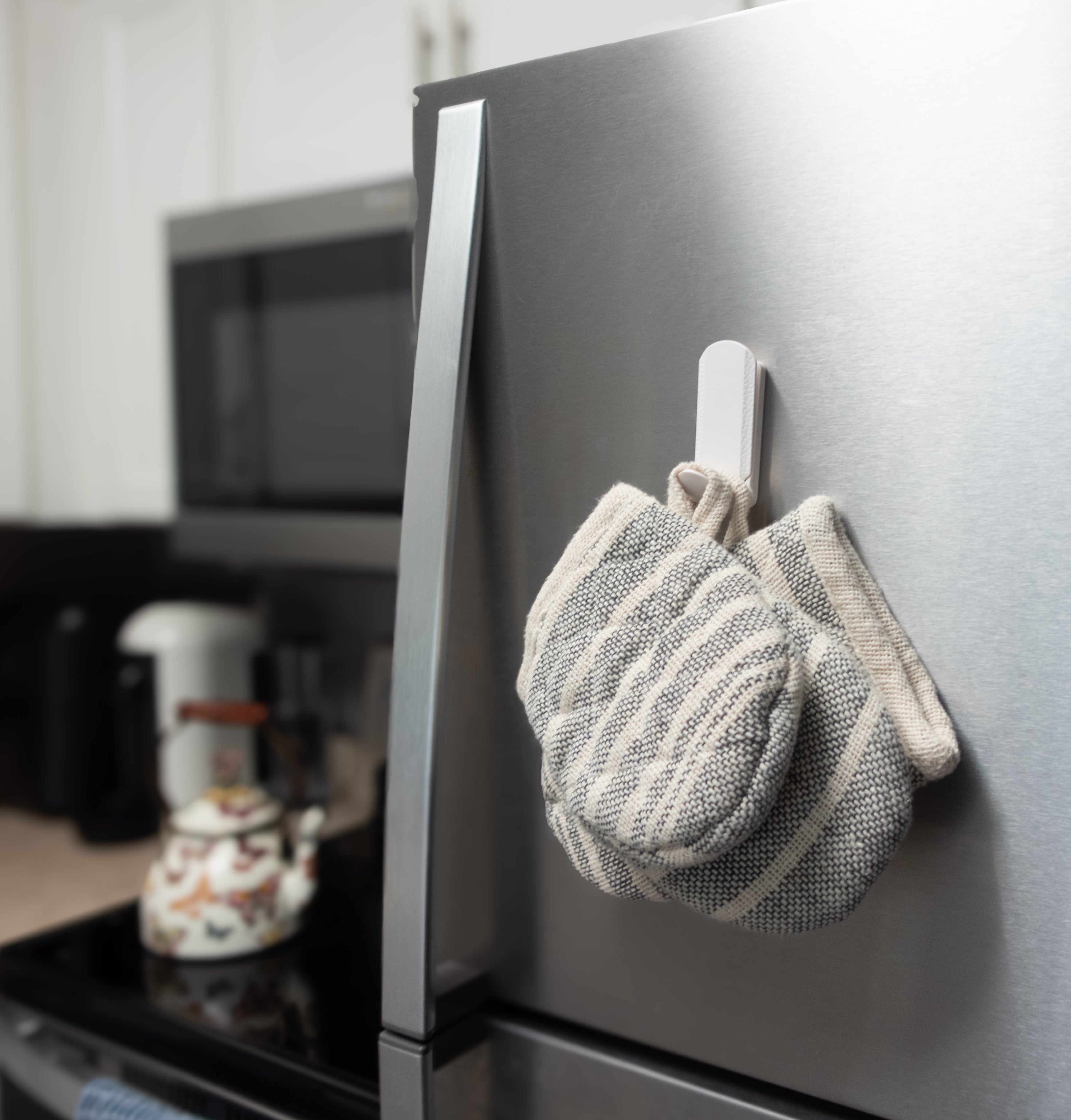 White magnetic hook attached to a stainless steel refrigerator holding a pair of striped oven mitts, demonstrating strong kitchen storage and organization.