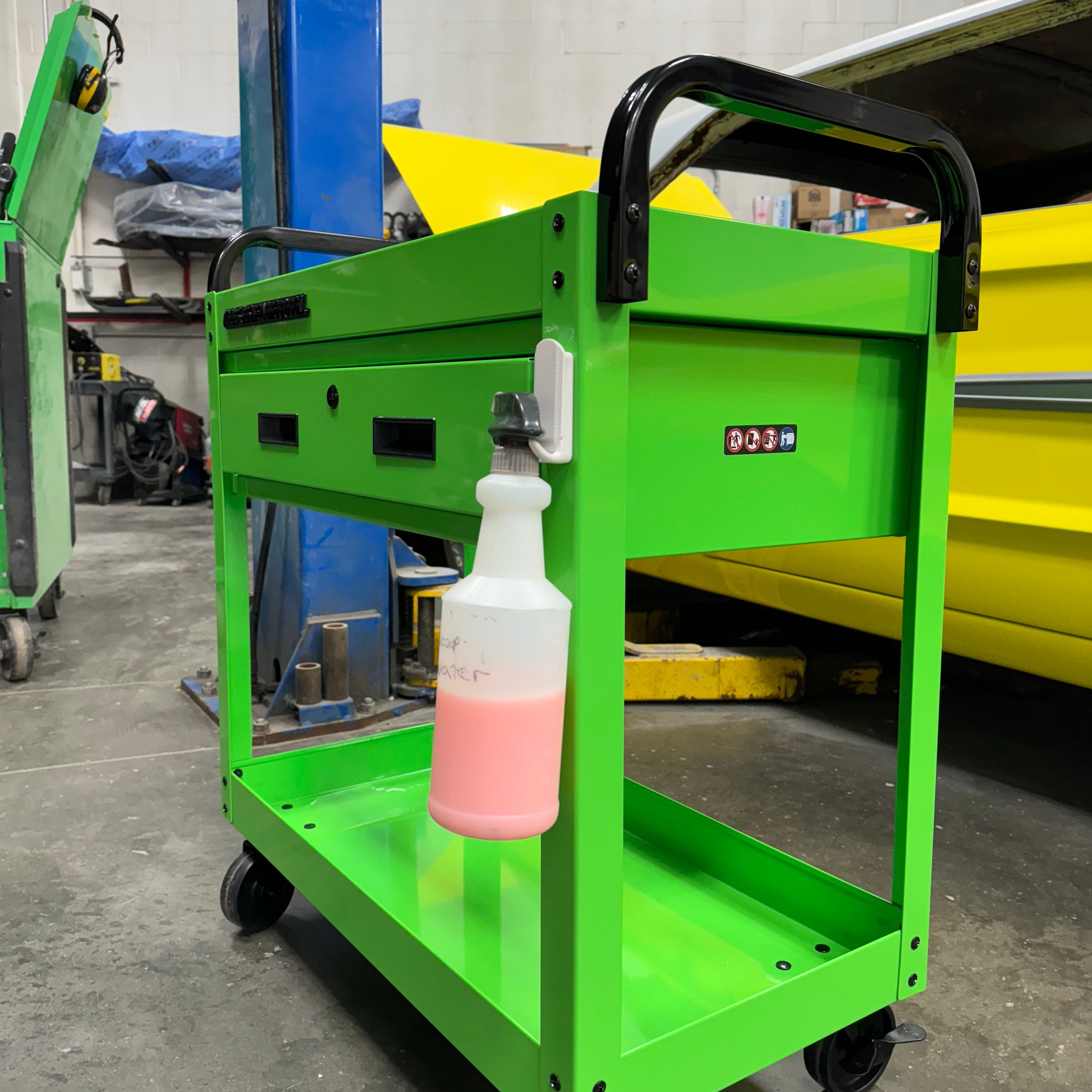 White magnetic hook attached to a bright green steel tool cart holding a spray bottle with pink liquid, showcasing convenient garage or workshop storage.
