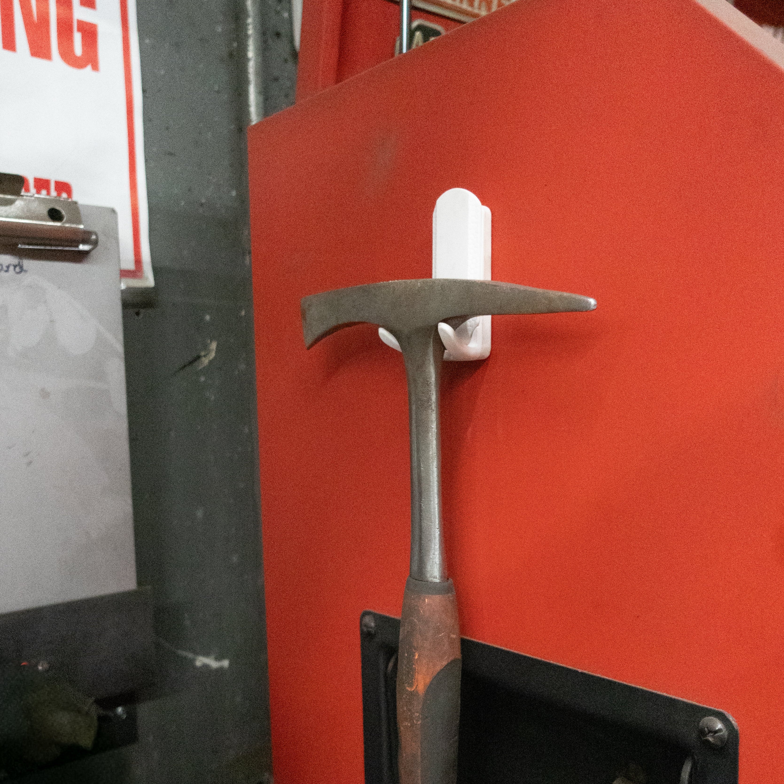 A rock hammer with a worn handle is hanging on a red steel toolbox side panel using a white Gator Magnetics magnetic hook, demonstrating efficient use of golf bag hooks for tool organization in a workshop.