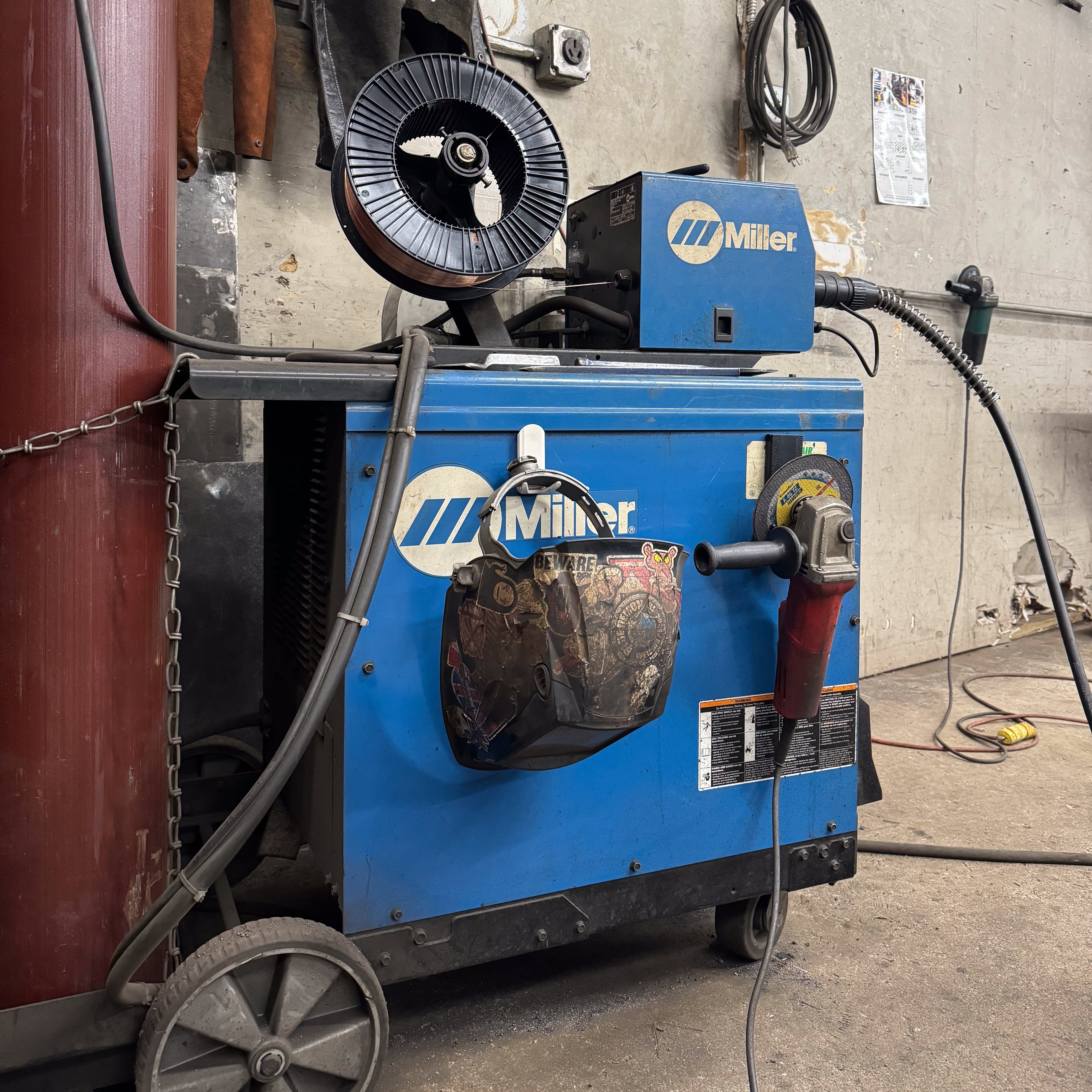 White magnetic hook attached to a blue Miller welding machine holding a welding helmet covered in stickers, demonstrating practical and strong storage in an industrial workspace.