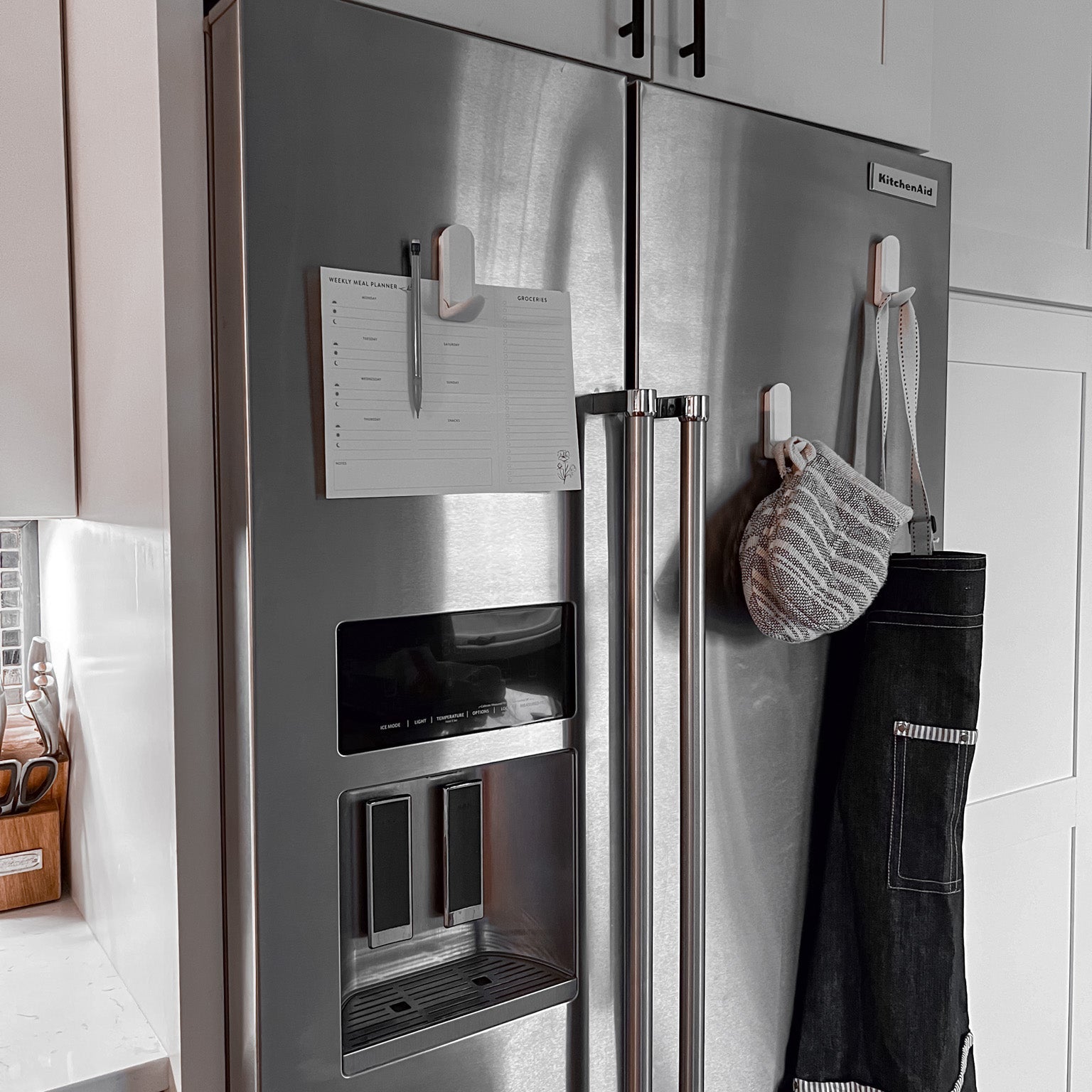White magnetic hooks on a stainless steel refrigerator holding a meal planning notepad with pen, a striped oven mitt, and a black kitchen apron, showcasing organized kitchen storage.