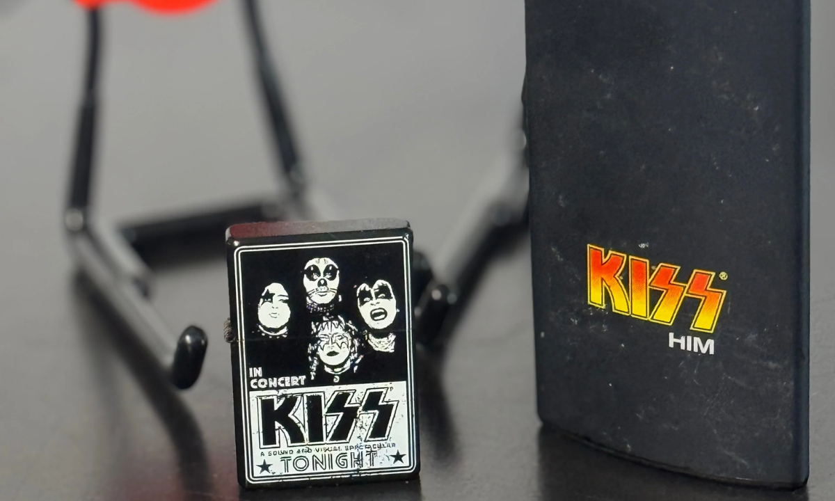 A foldable guitar stand and a black flip phone with colorful "KISS HIM" text sit on a flat black table surface.