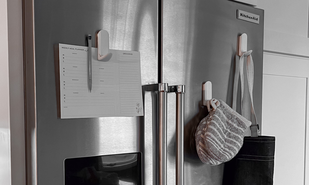Three white Gator magnetic hooks hold a meal planner and oven mitt on a stainless steel refrigerator.