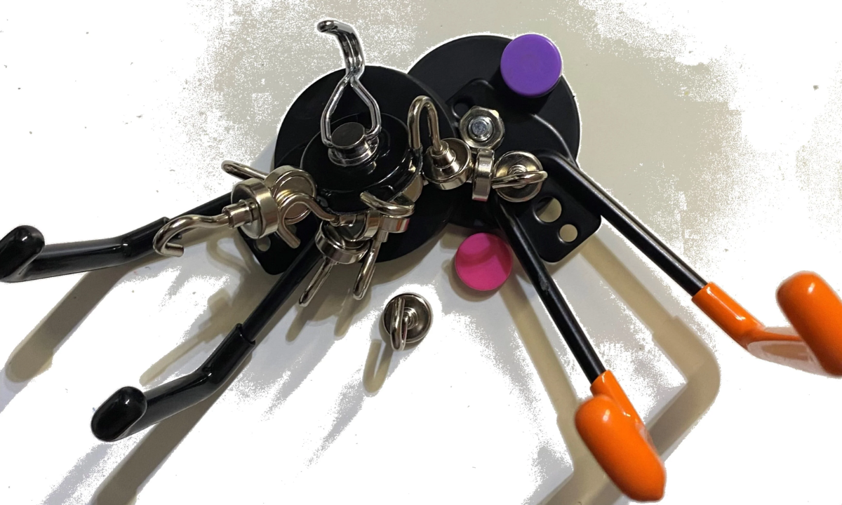 Two Gator magnetic hooks hold key rings and carabiners on a steel wall beside magnetic push pins.