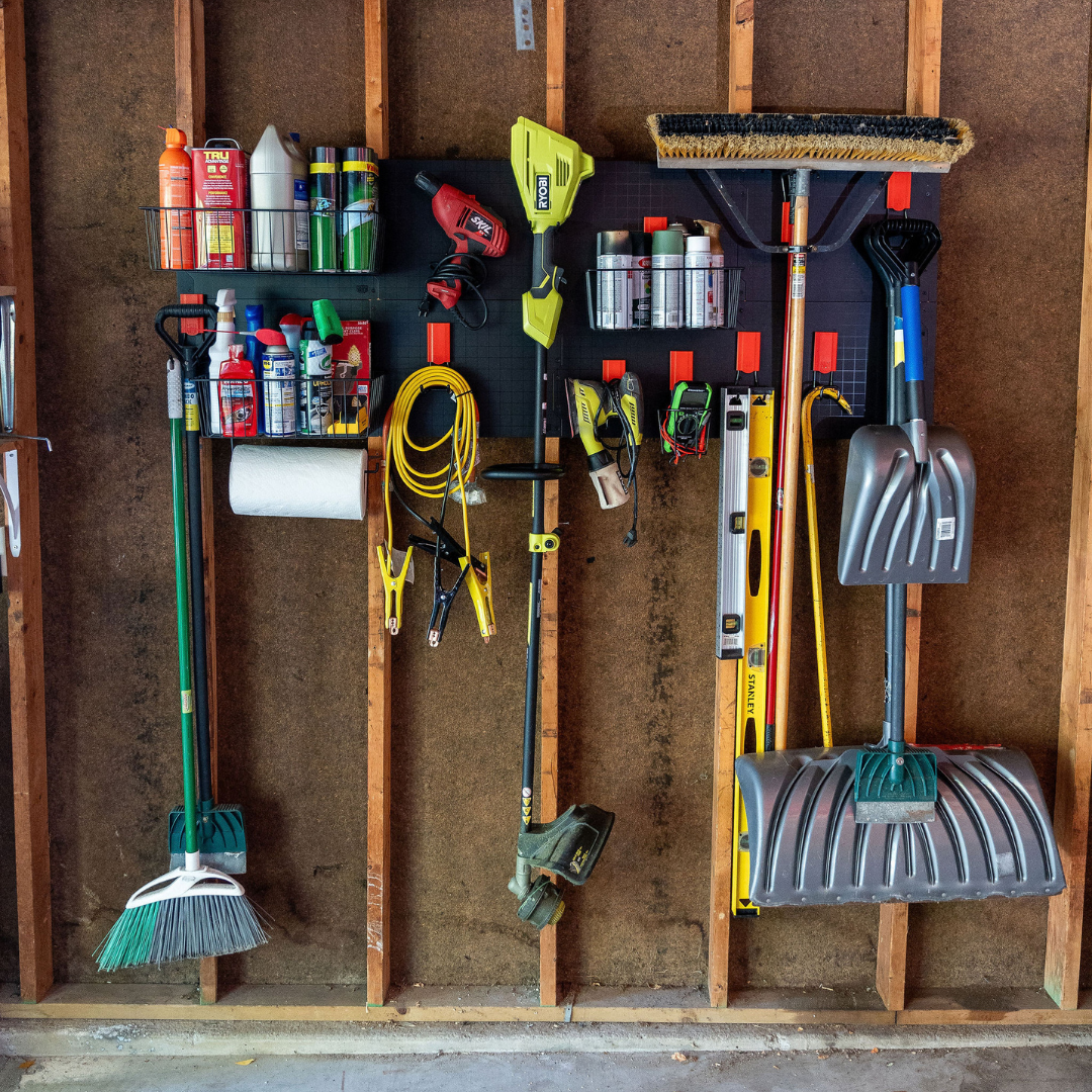 A black Gator Steel Wall Panel system mounted on a pole barn steel wall holds red Gator Magnetics hooks and magnetic storage baskets storing spray bottles, aerosol cans, paper towels, an extension cord, a heat gun, a string trimmer, a level, a rake, a broom, a shovel, jumper cables, a measuring tape, and a snow shovel.