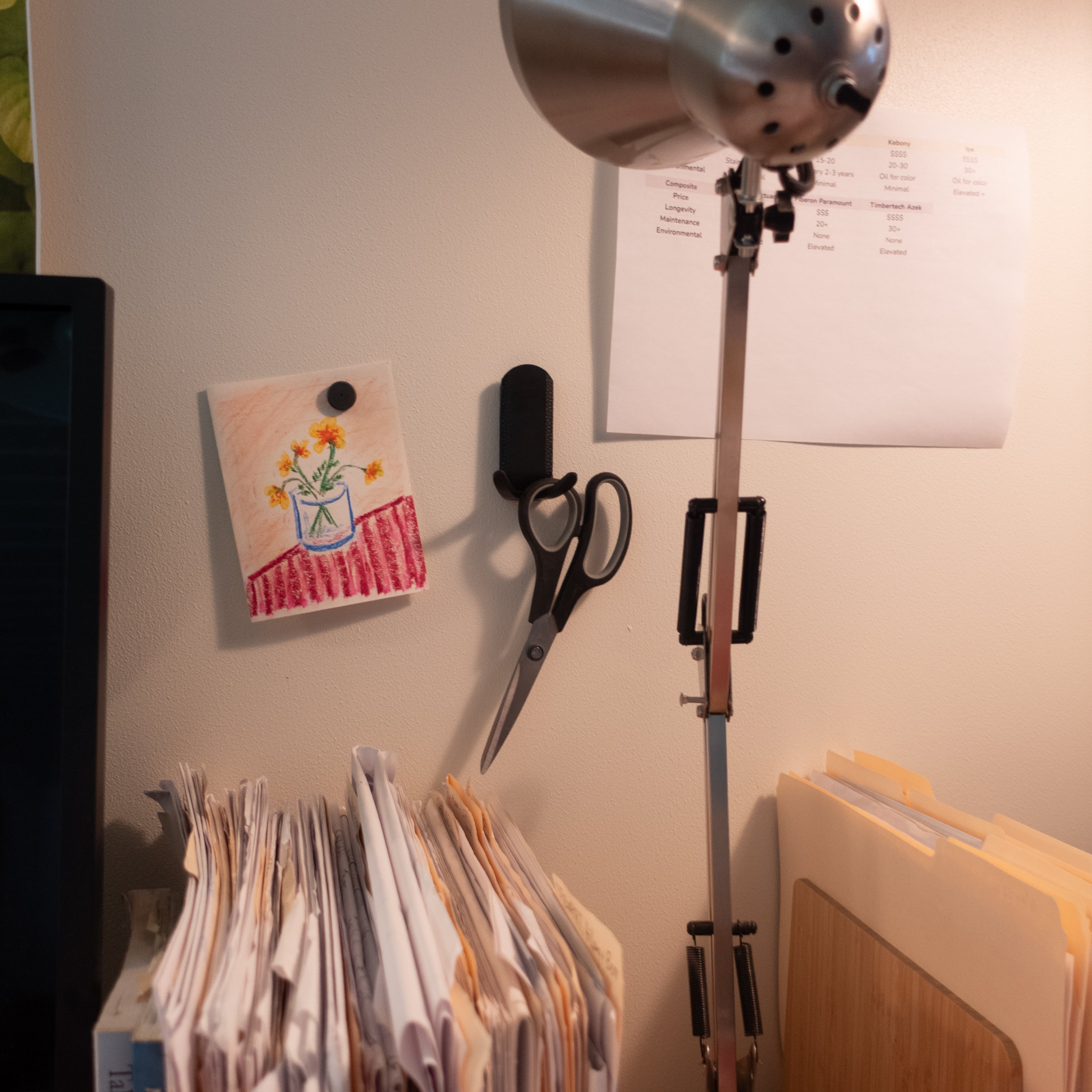Black magnetic hook on a steel wall in a home office holding a pair of scissors next to a desk lamp, with a small drawing and paperwork nearby, showcasing organized workspace storage.