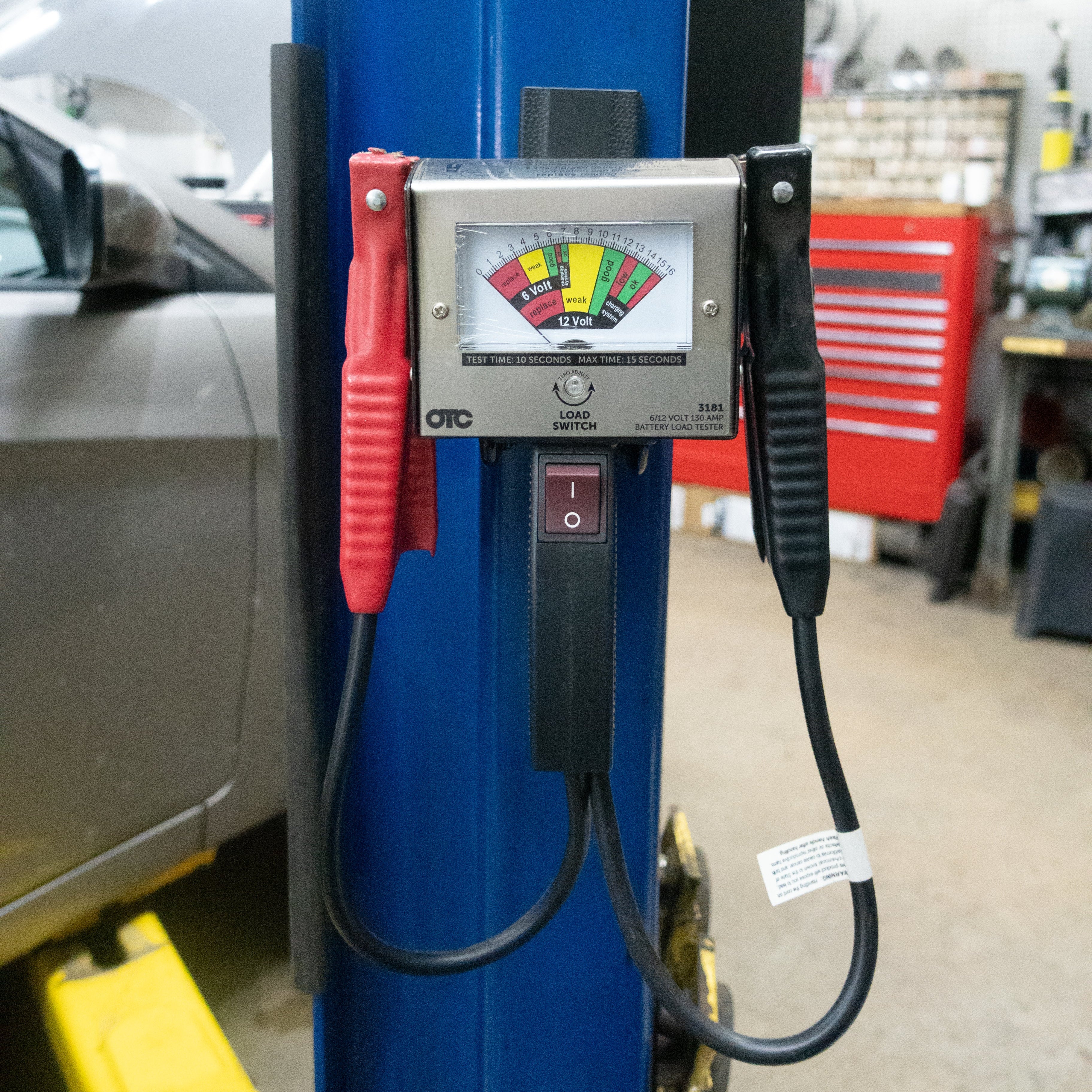 OTC battery load tester with cables hanging from a Gator Magnetics hook mounted on a blue automotive lift in a repair shop