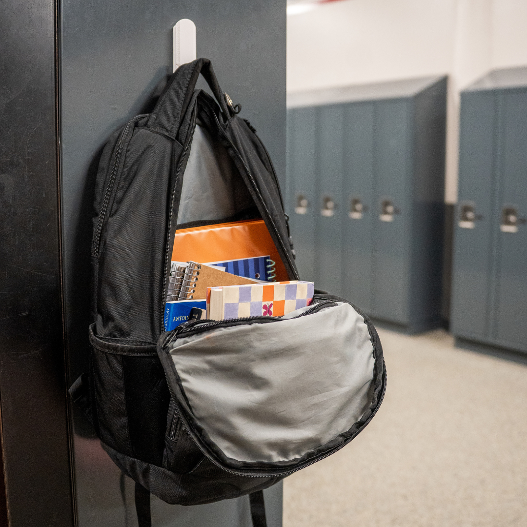 Black backpack filled with notebooks hanging from a white Gator magnetic hook on a steel locker door.