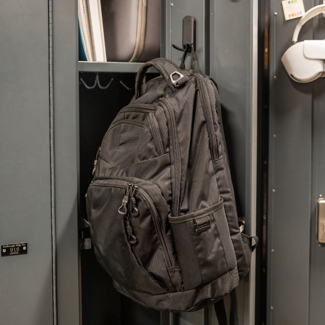 A black backpack is hung by its strap on a red Gator Magnetics magnetic hook attached to a steel locker interior panel.