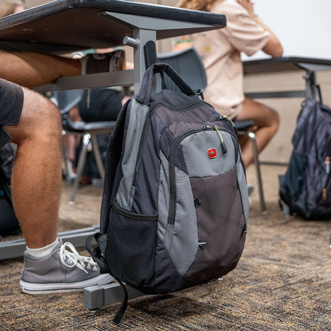 Black and gray backpack hanging from a Gator magnetic hook on a steel classroom desk.