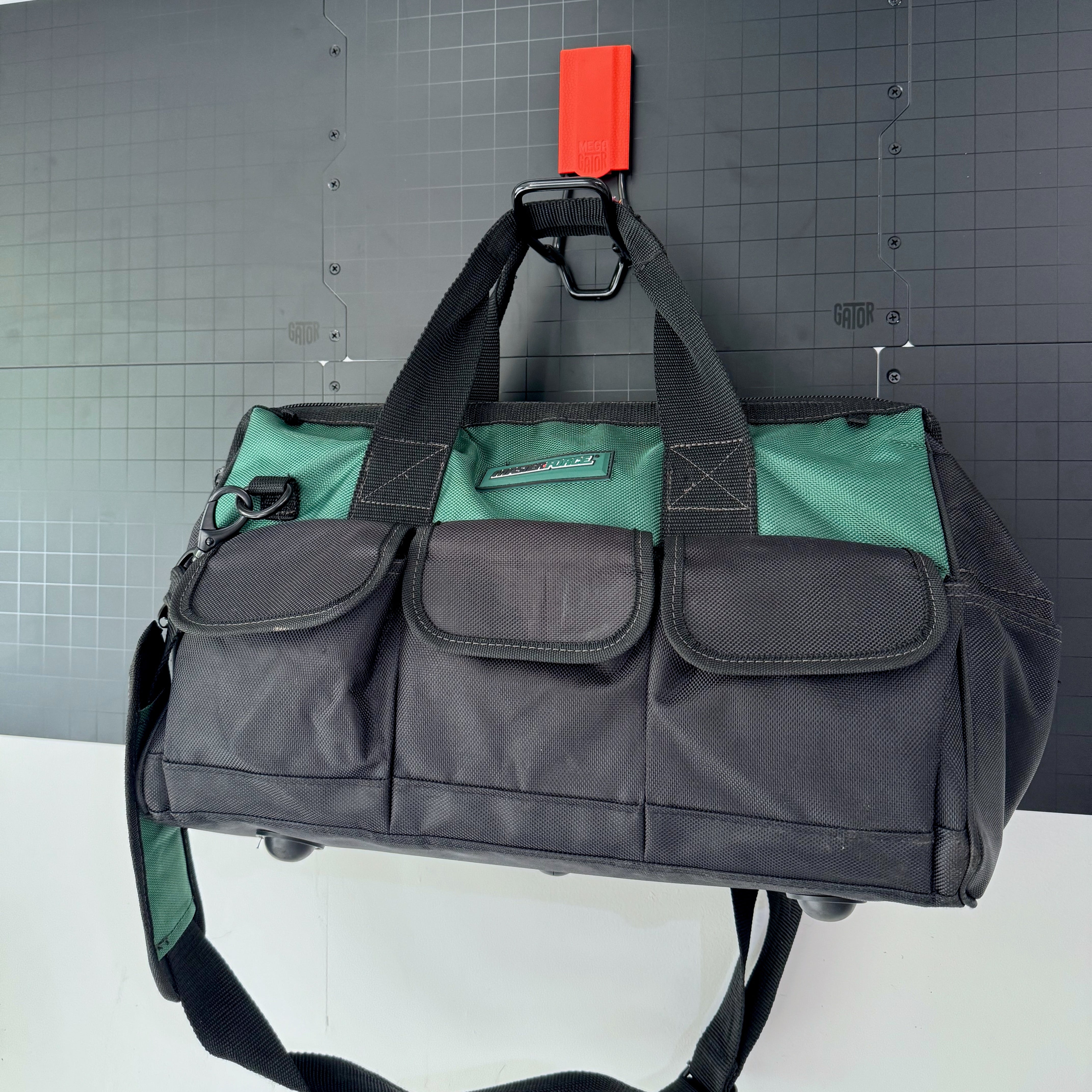 A black and green tool bag is suspended by its handle on a red Gator Magnetics magnetic hook attached to a black Gator Steel Wall Panel.
