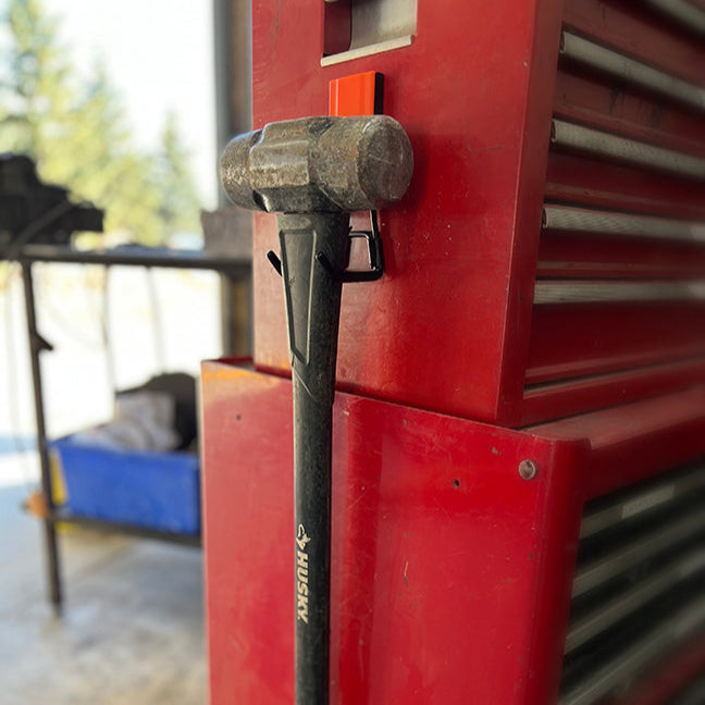 A black-handled sledgehammer with a worn metal head is hanging on a red steel toolbox side panel using a Gator Magnetics red magnetic hook.