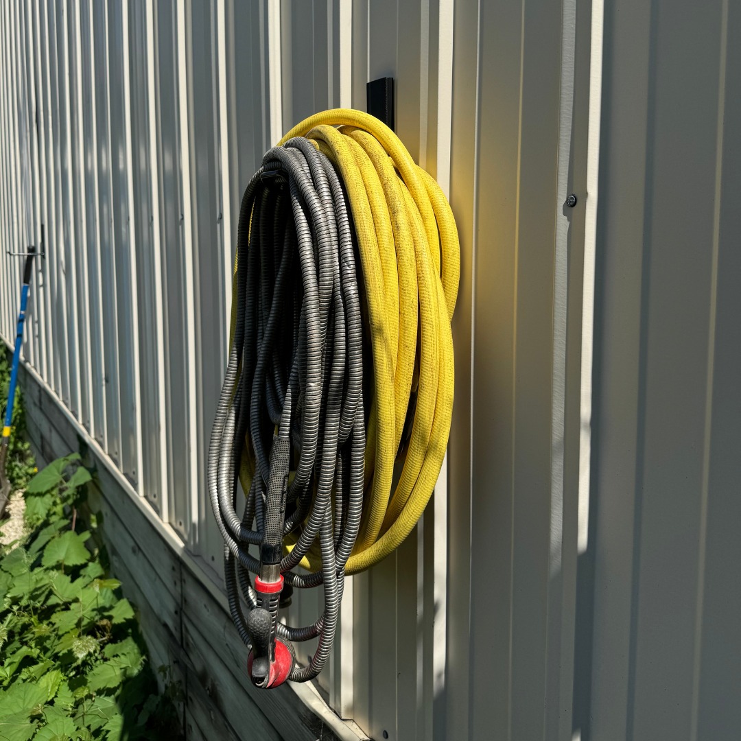 A black magnetic hook from Gator Magnetics holds coiled black and yellow garden hoses on a vertical corrugated steel wall.