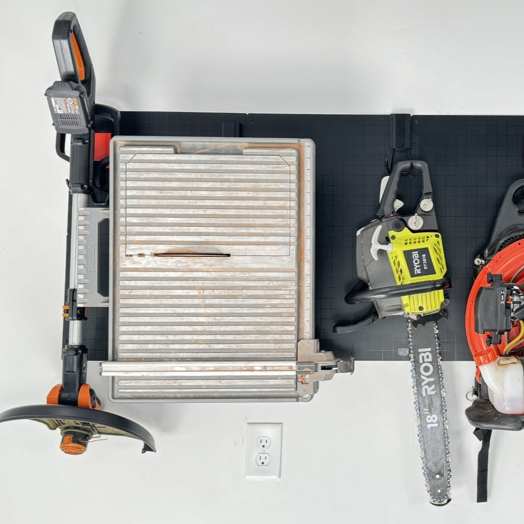 A black Gator Steel Wall Panel in a garage holds a Ryobi chainsaw by the bar and a metal tile cutter by its handle.