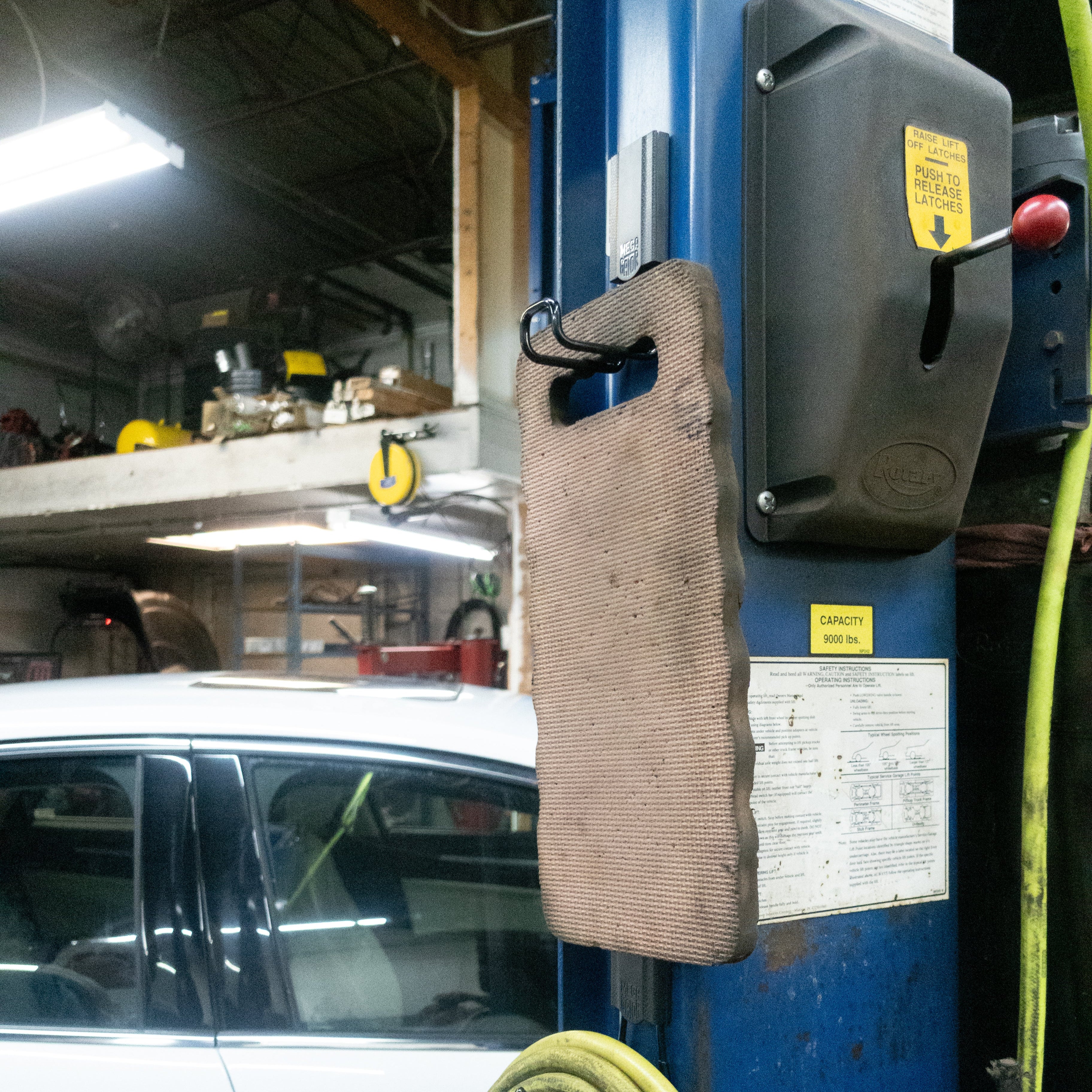 Brown kneeling pad hanging from two red Gator heavy-duty magnetic hooks on a blue steel vehicle lift.