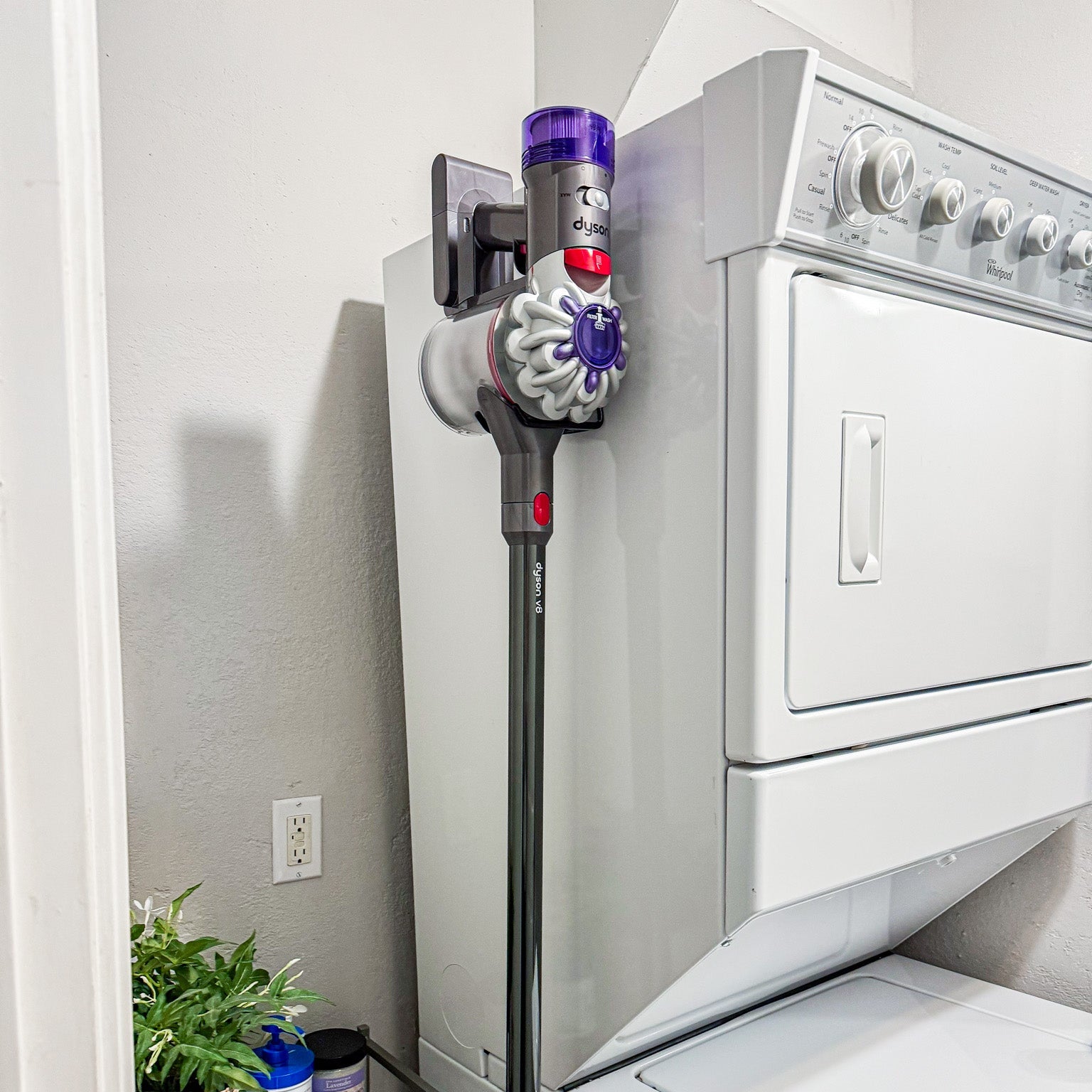 Cordless stick vacuum mounted on a wall garage steel panel adjacent to a stacked washer and dryer in a laundry room.
