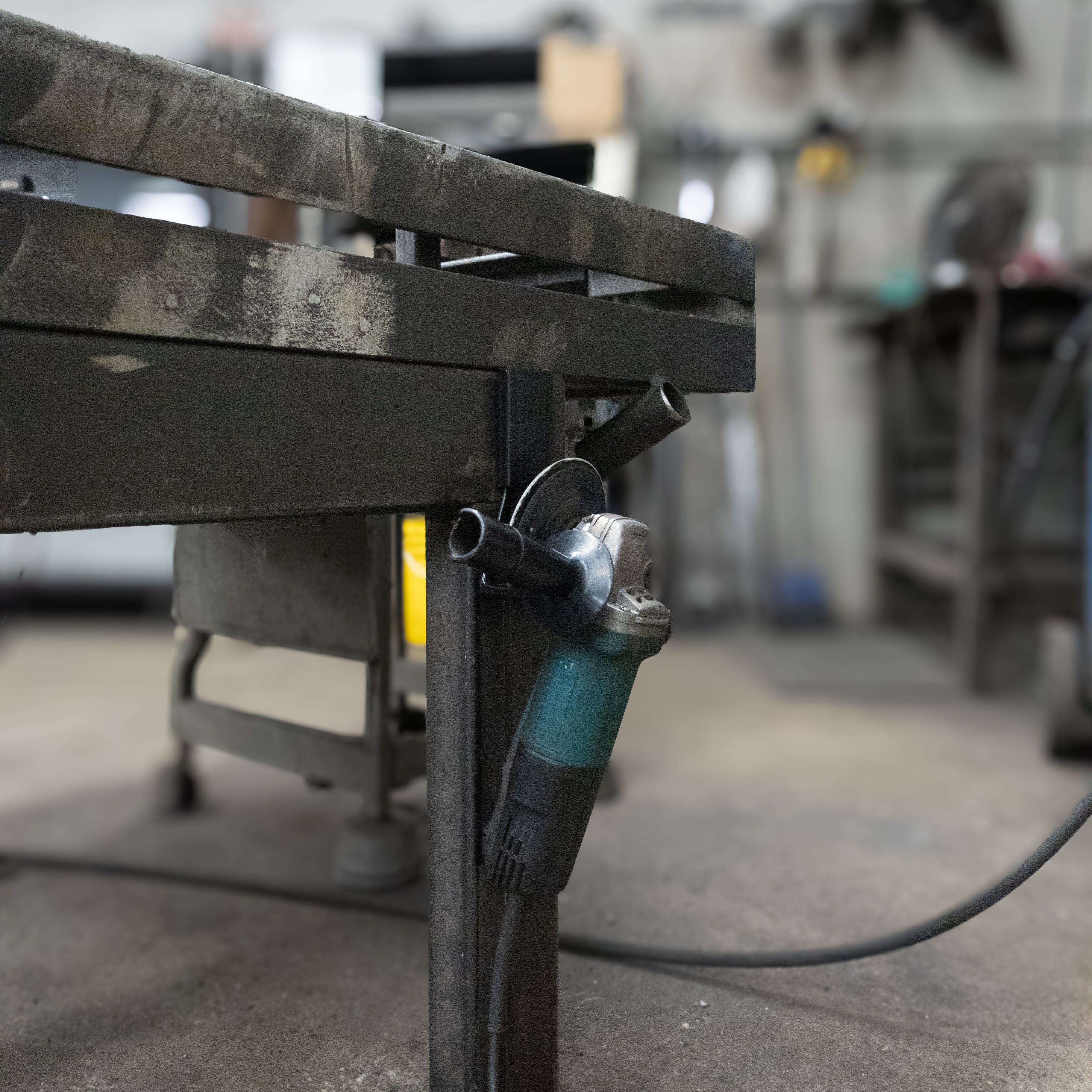 An electric angle grinder is suspended by a red Gator Magnetics hook on the leg of a heavy-duty steel workbench in a workshop.