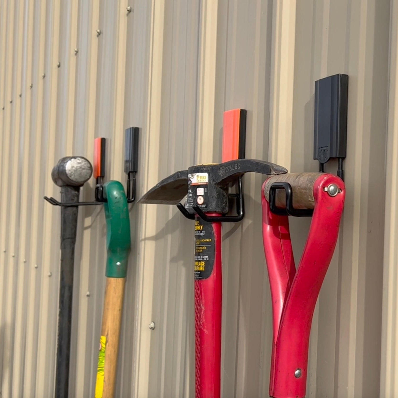 Four red Gator magnetic hooks hold a sledgehammer, garden tool, pickaxe and bolt cutter on corrugated steel.