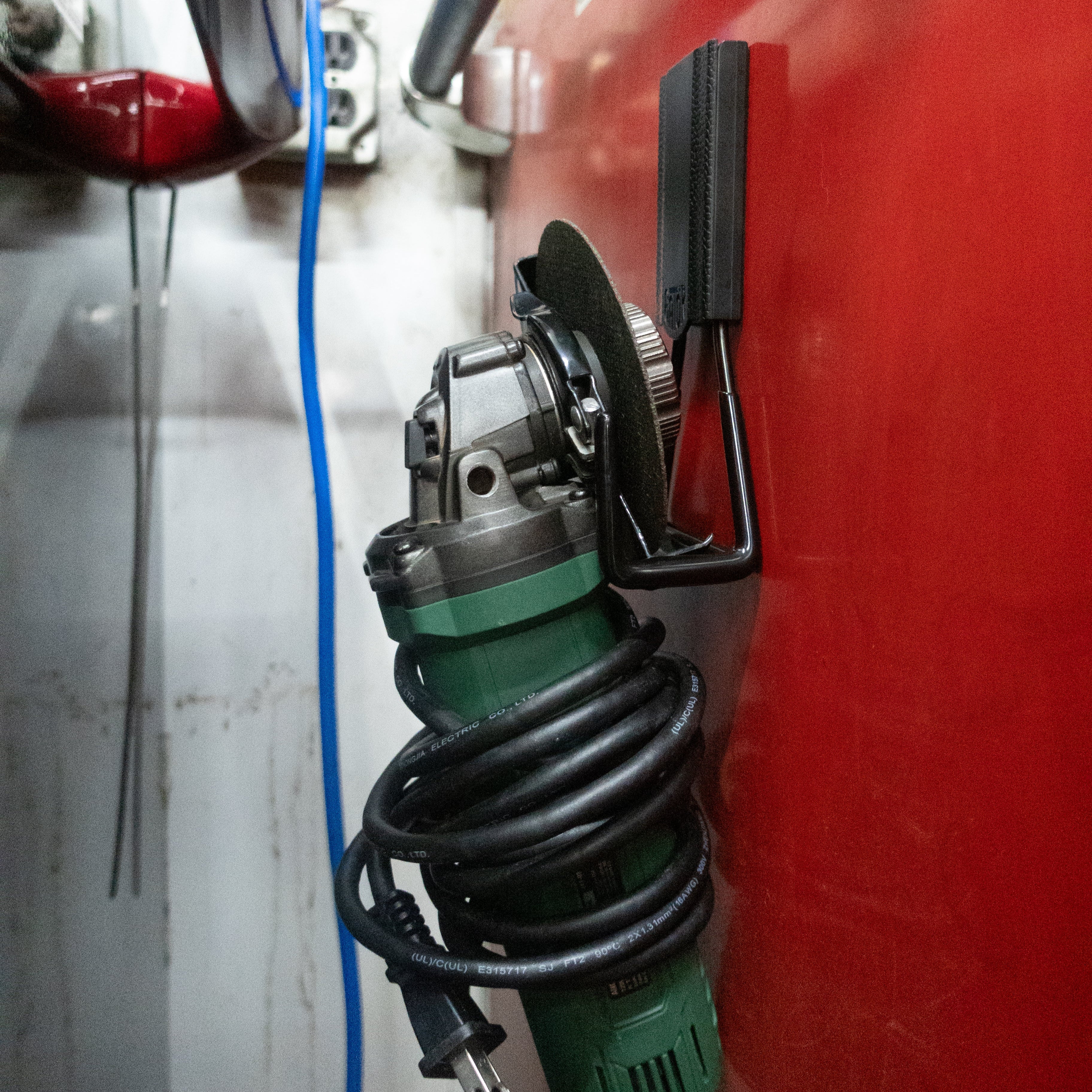 Green angle grinder with coiled cord hanging from a black Gator magnetic hook on a red steel wall panel.