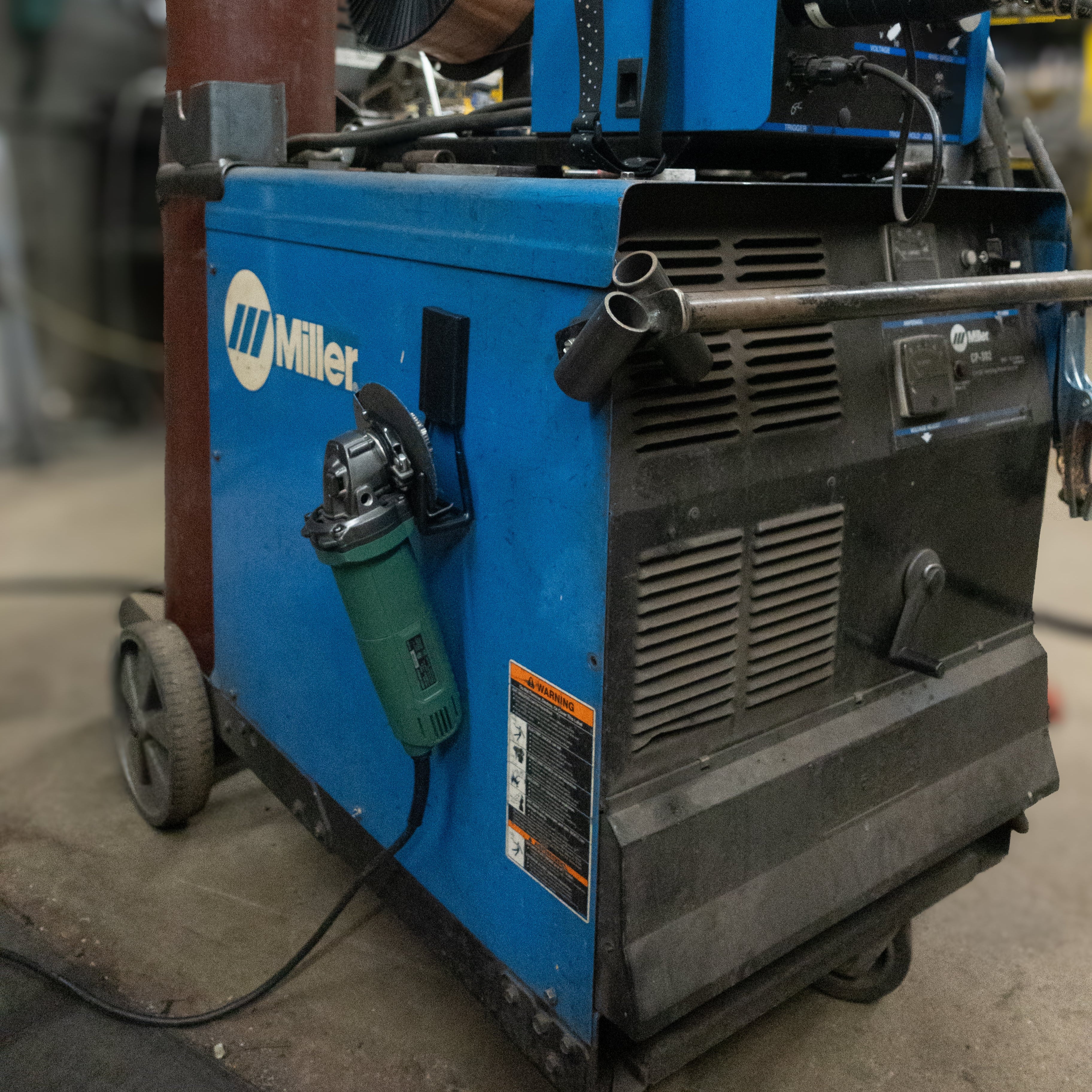 A green angle grinder is hanging by its handle on a red magnetic hook attached to the blue steel side panel of a Miller welding machine.