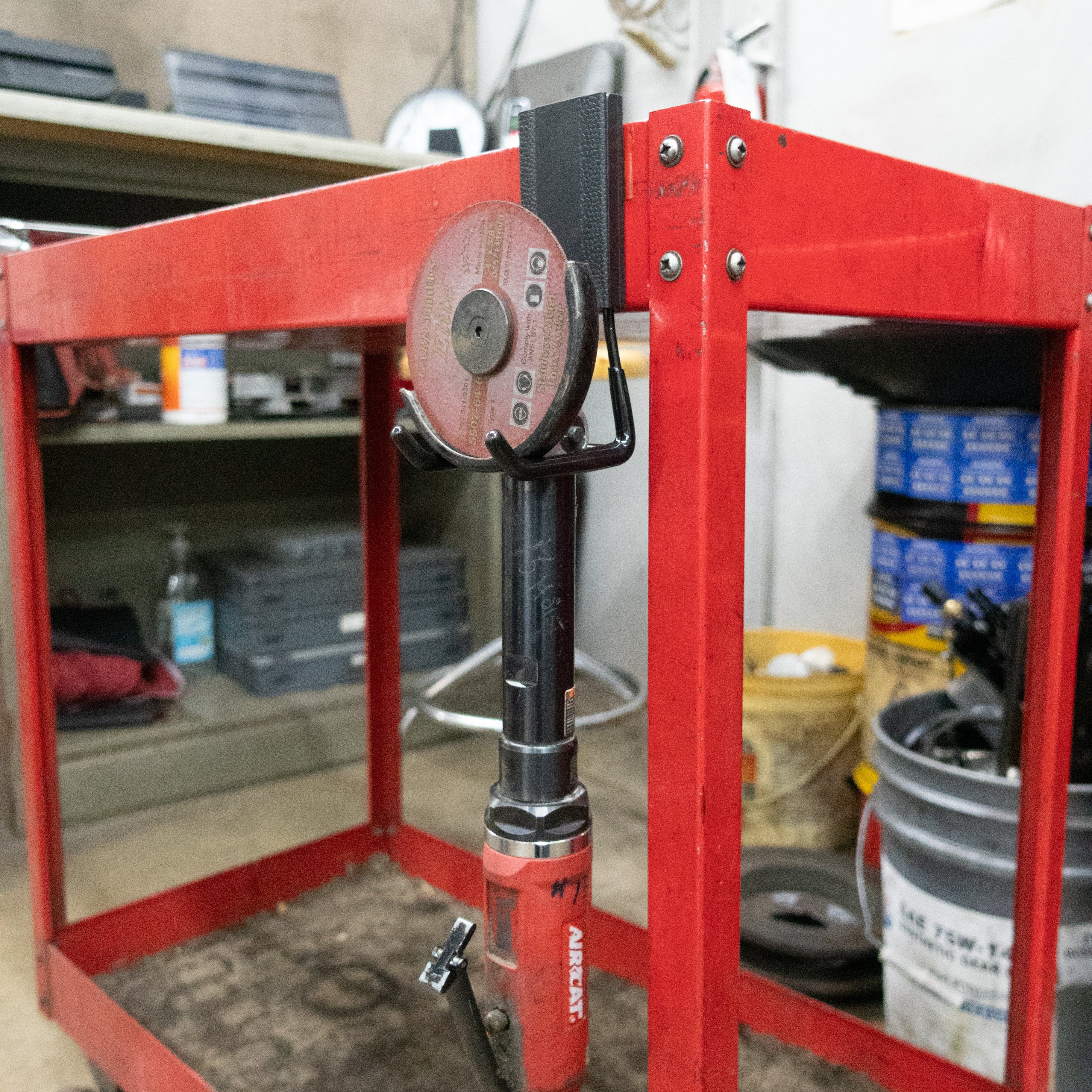 Red angle grinder with cutting disc securely hung on a Gator Magnetics hook mounted to a red metal workbench frame in a workshop