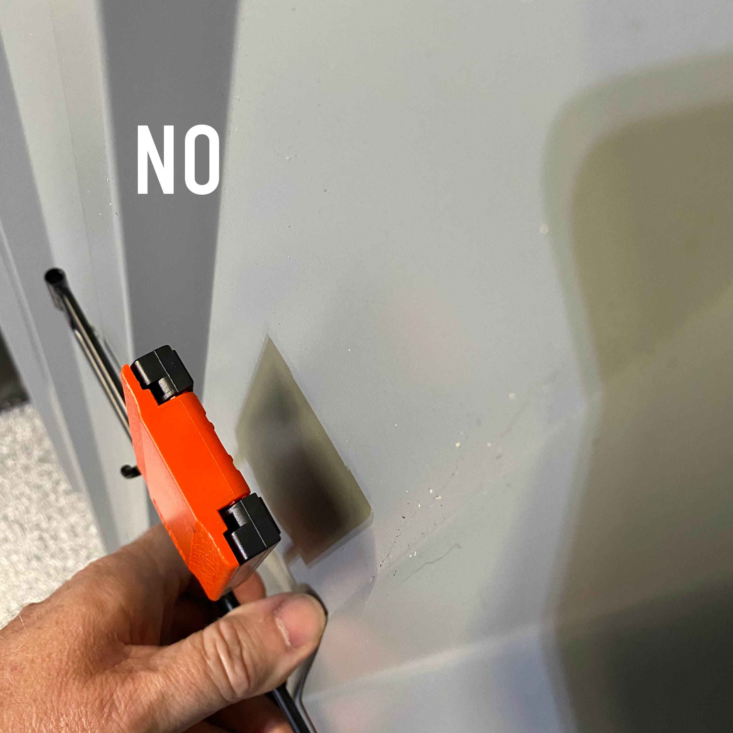 A hand holds a red magnetic hook near a smooth gray painted steel surface with the word "NO" visible.