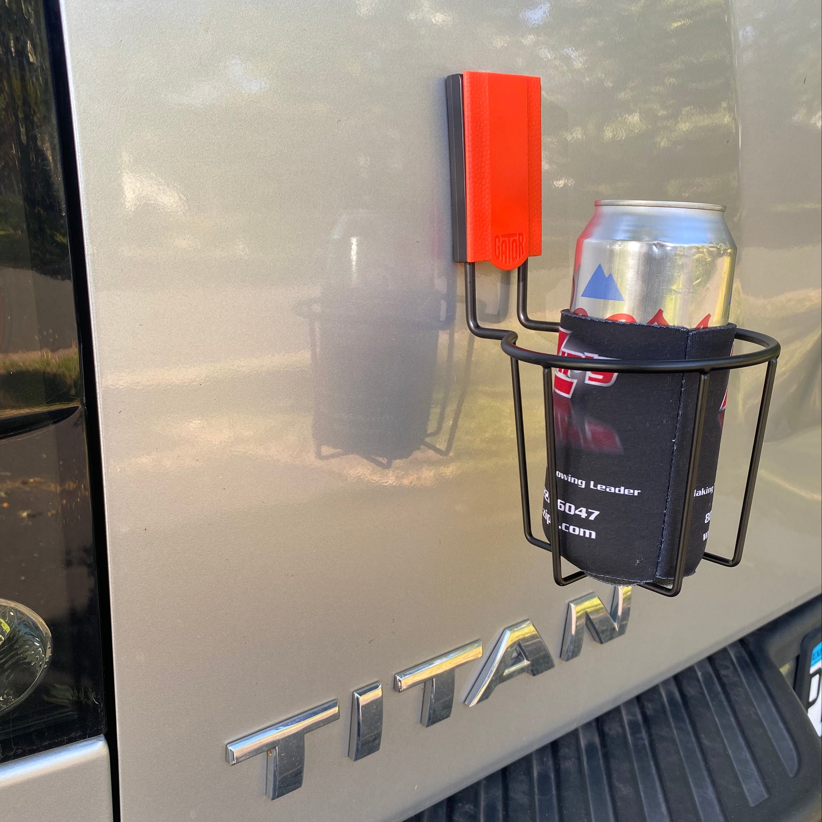A Gator Magnetics magnetic cup holder is attached to the steel tailgate of a truck, holding a canned drink wrapped in a black koozie.