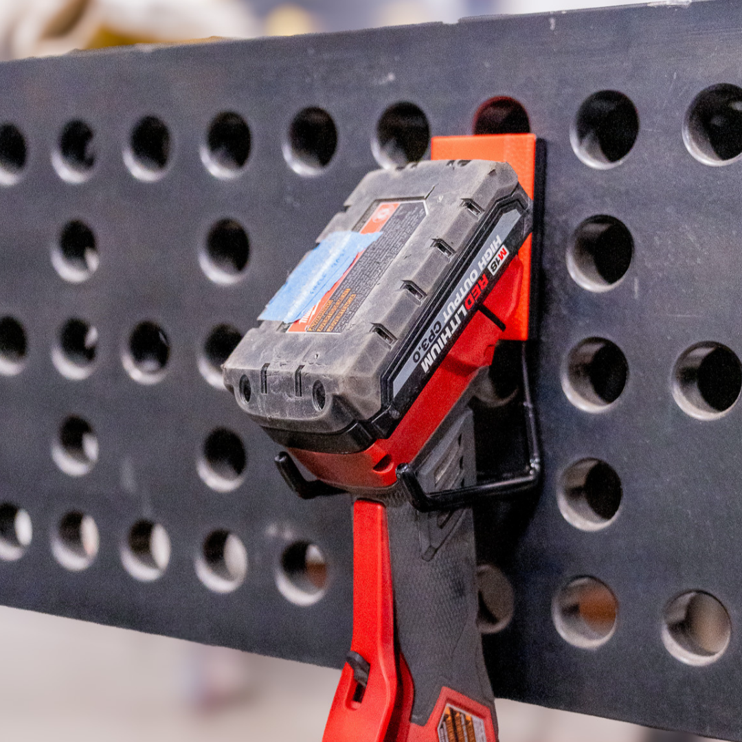 Milwaukee power tool battery held by a red Gator magnetic hook on a perforated black steel tool board.