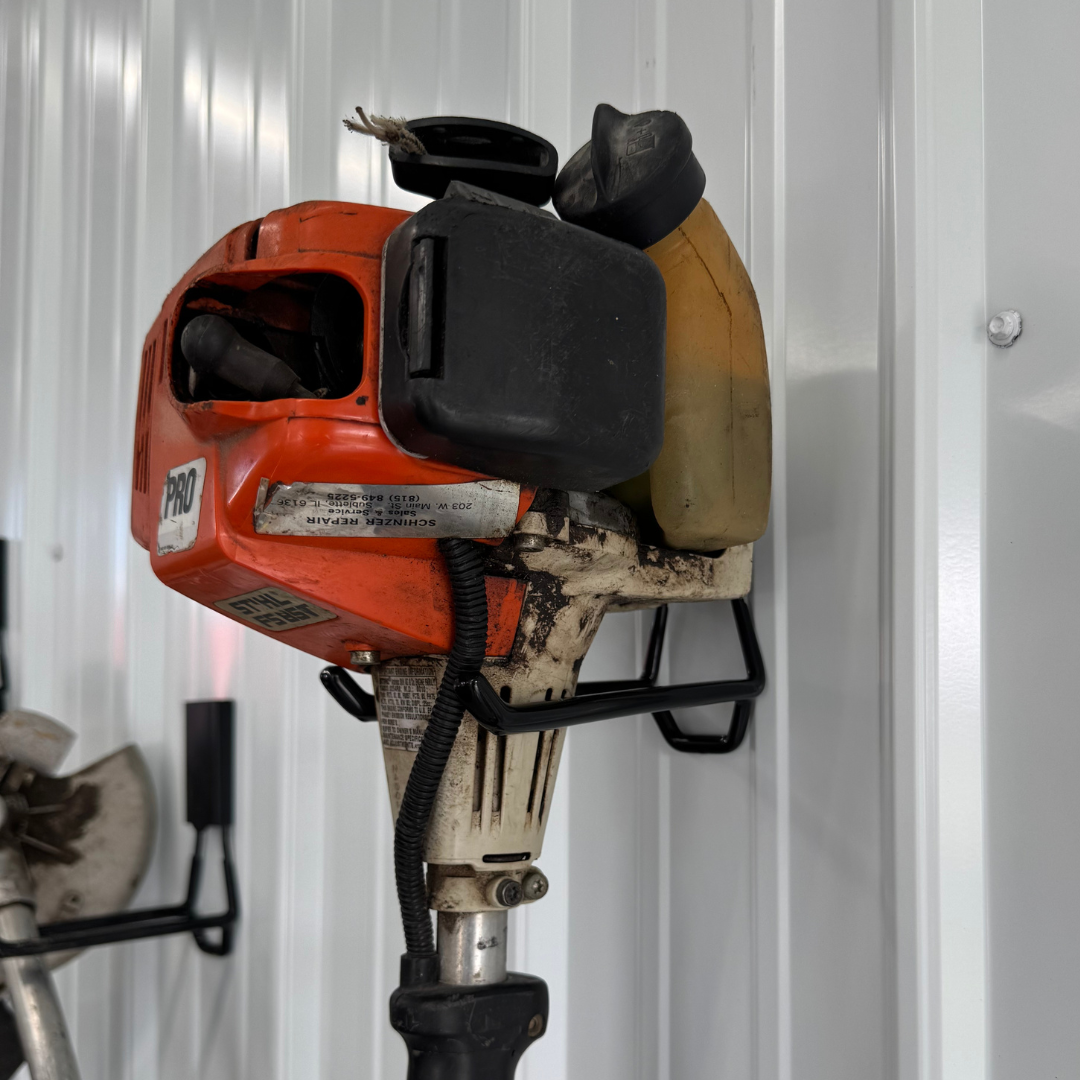 An orange and white gas-powered trimmer is suspended by a black magnetic hook on a vertical steel wall panel.