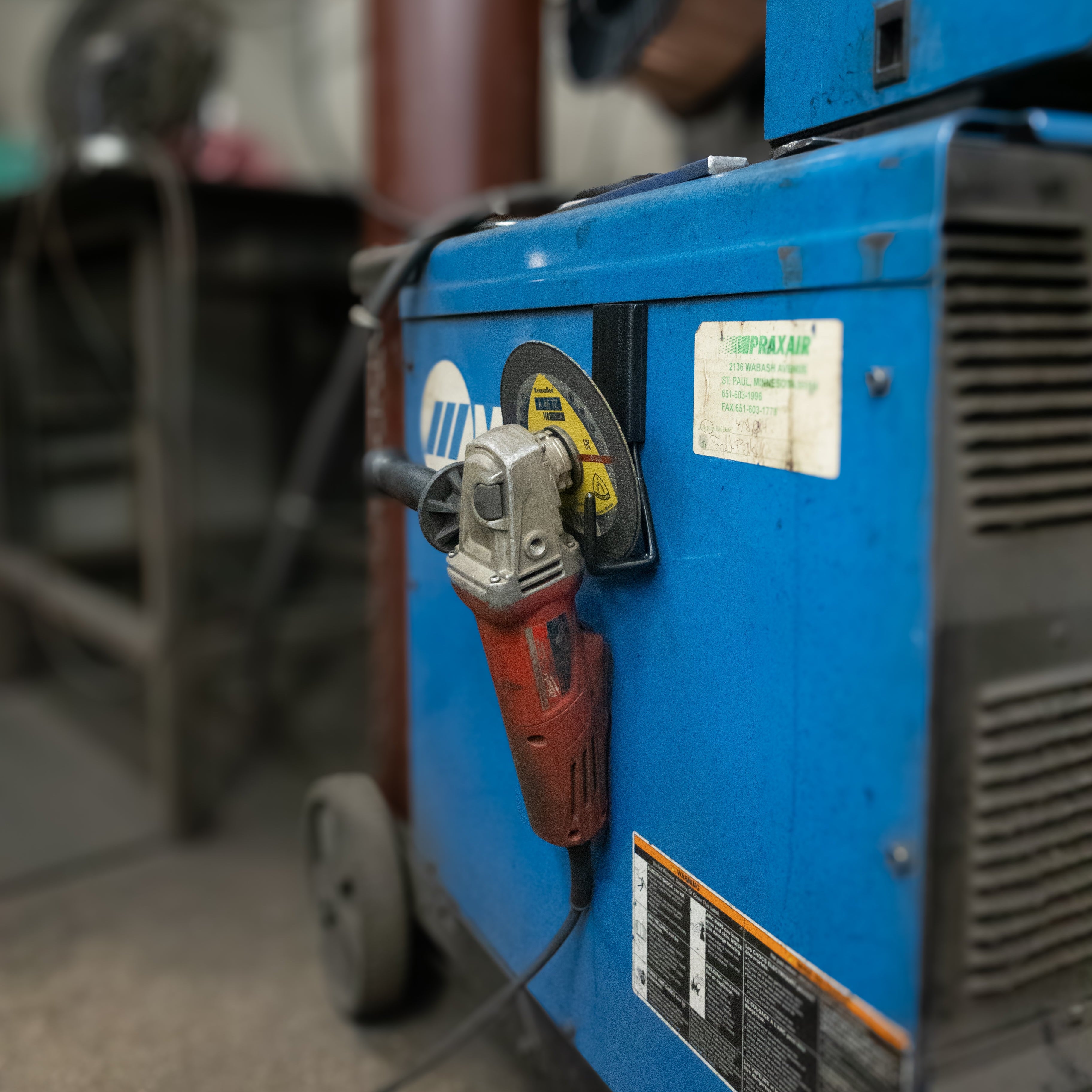 Red angle grinder hanging from a black Gator magnetic hook on a blue steel welding machine.