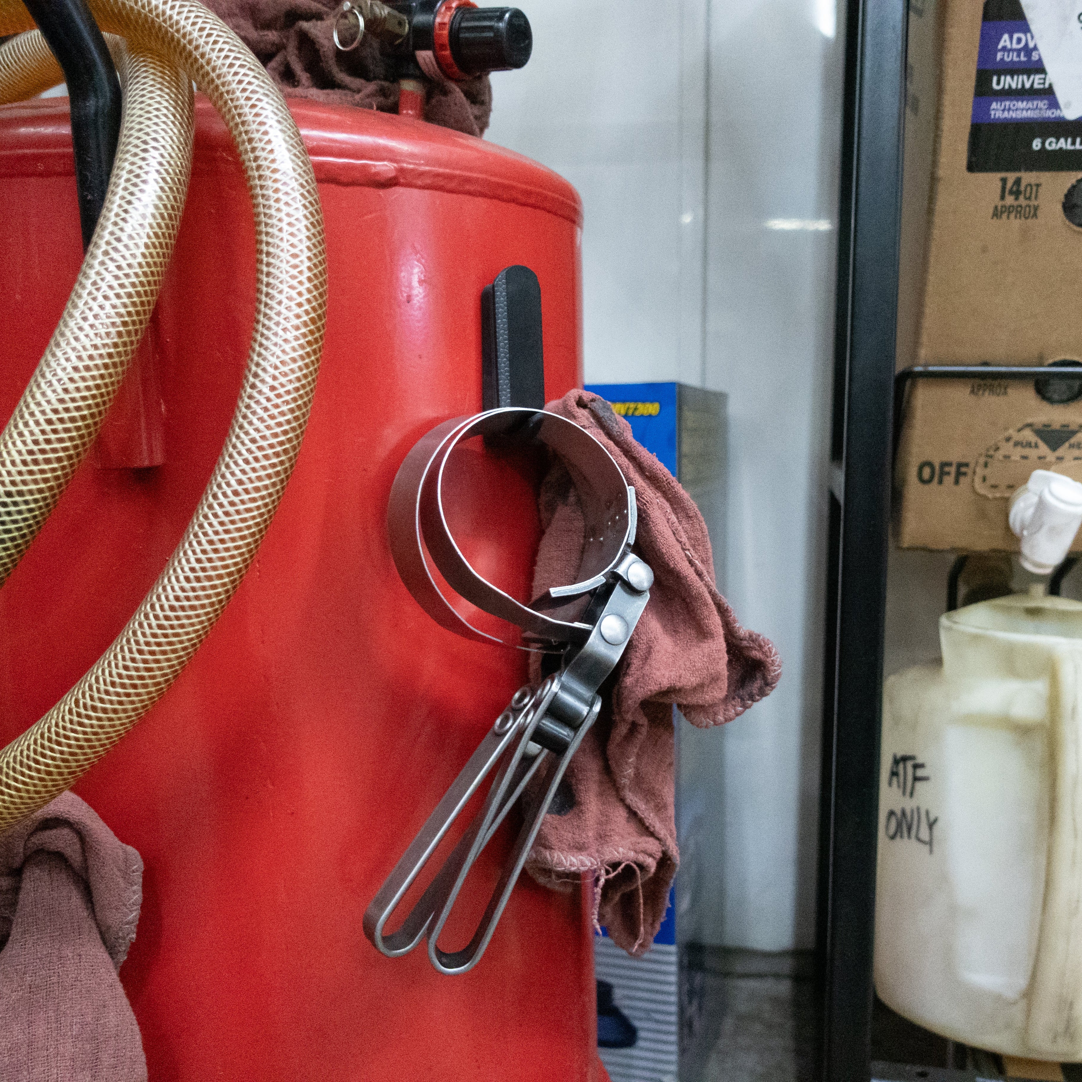 A red Gator Magnetics magnetic hook is attached to a red steel air compressor tank, holding a metal oil filter wrench and a brown shop rag.
