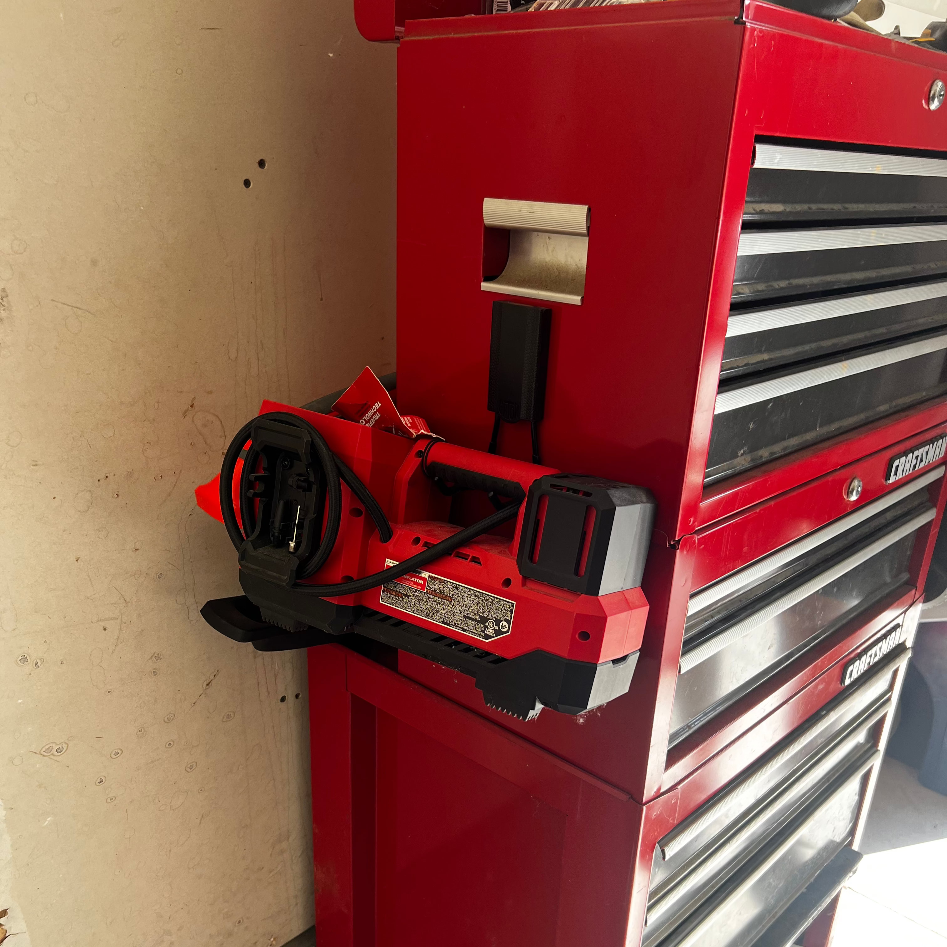 A red power tool battery charger is hung by a Gator Magnetics magnetic hook on the steel side panel of a red Craftsman tool chest.