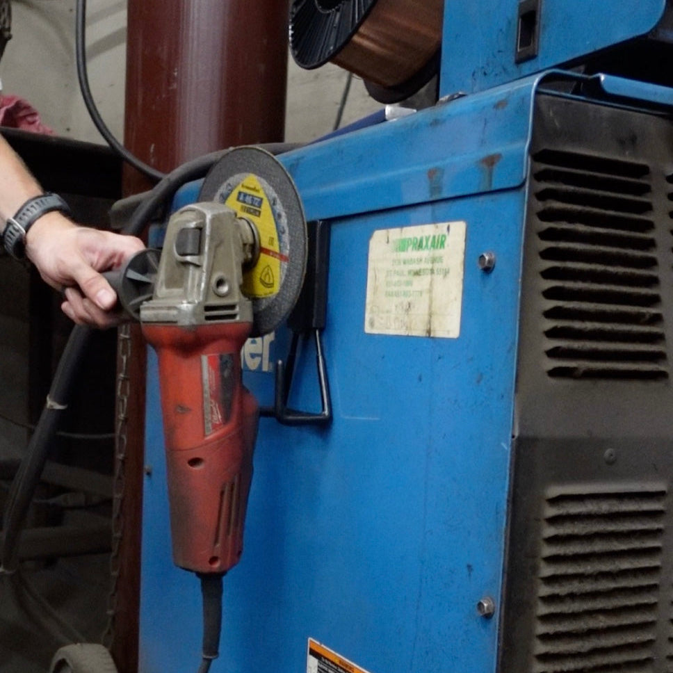 A red power tool grinder is hung by its handle on a black magnetic hook attached to a blue steel panel of industrial equipment.