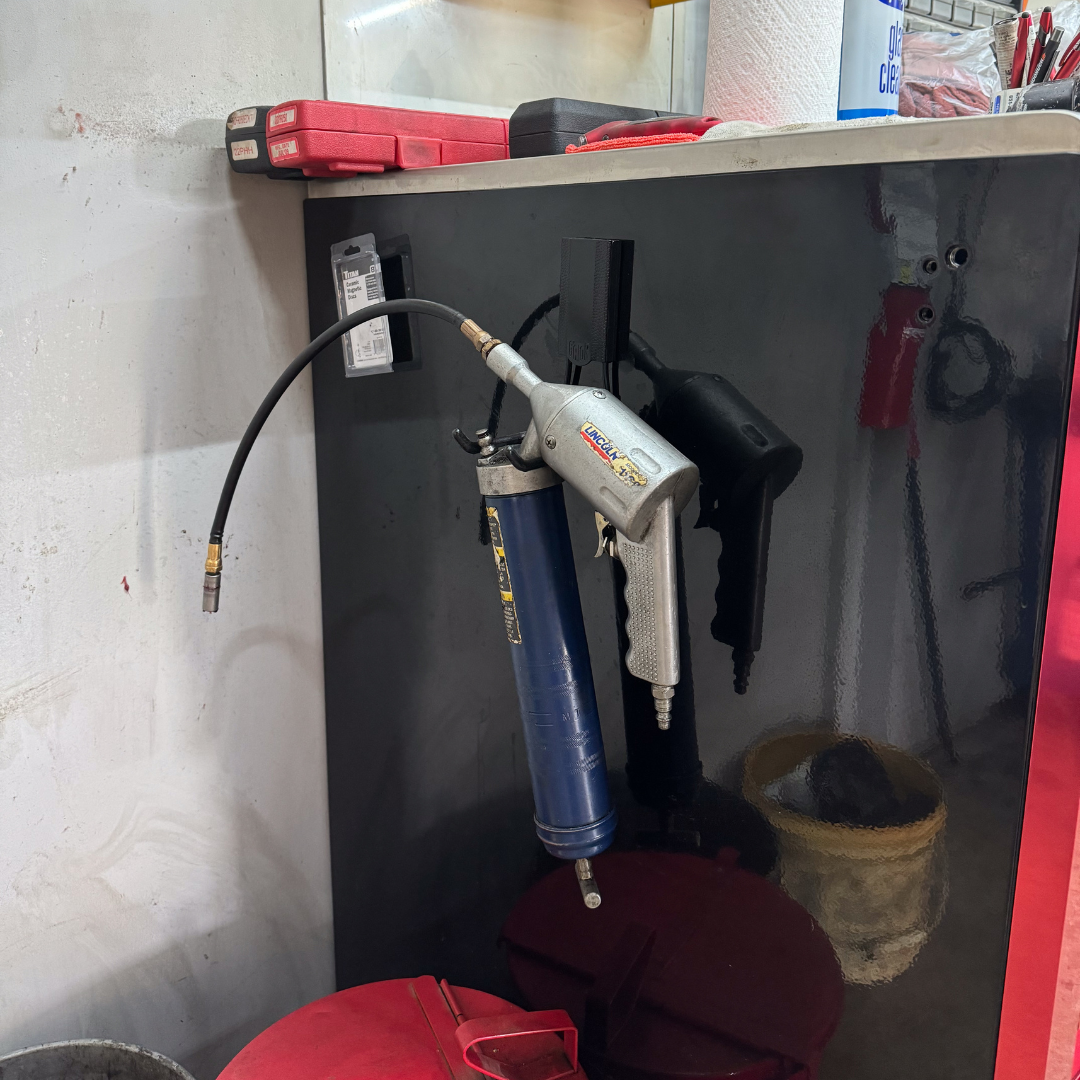 A silver pneumatic grease gun with an attached hose is hanging from a black steel toolbox side panel using a Gator Magnetics magnetic hook.