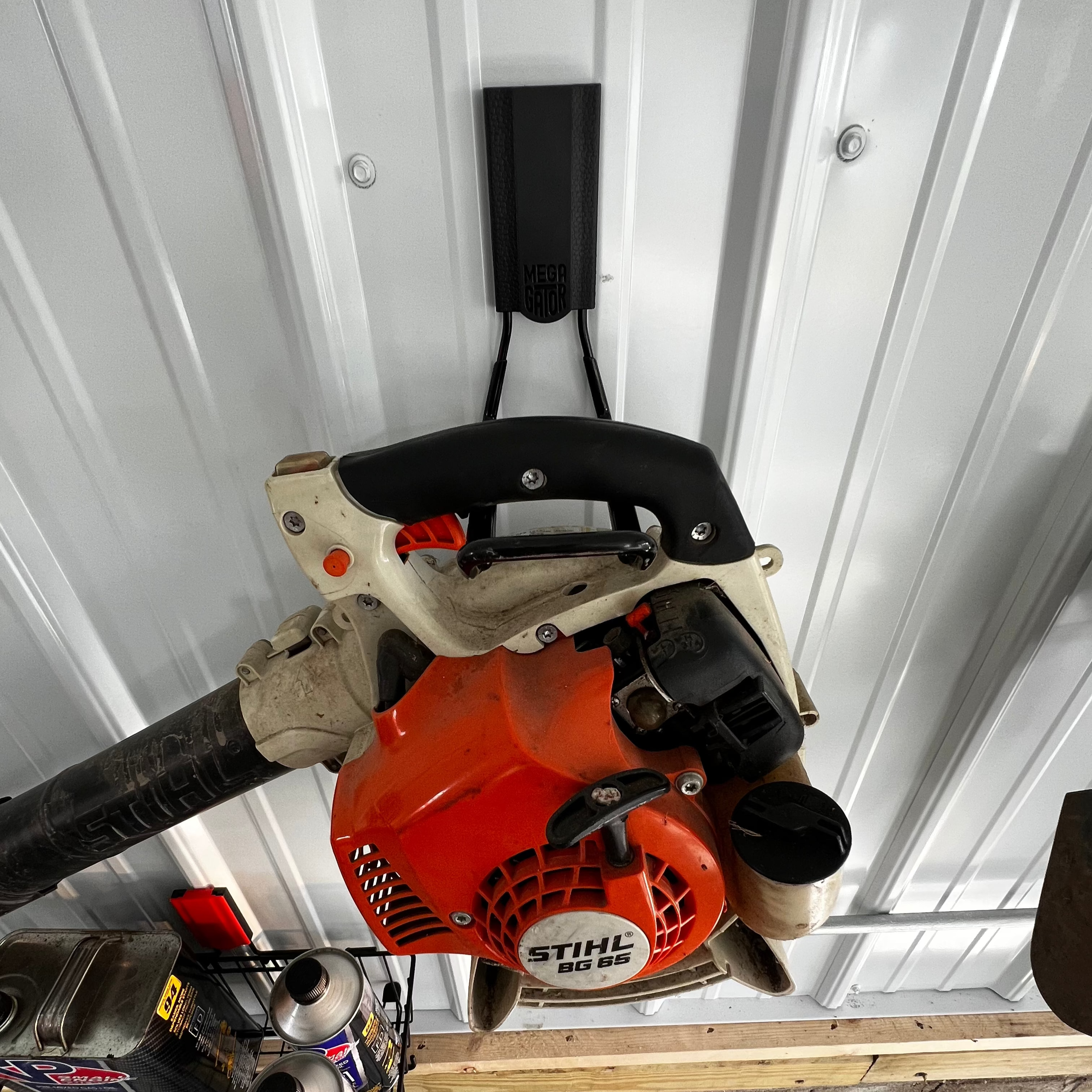 A Stihl BG 65 leaf blower is hung by its handle on a black Gator Magnetics hanger mounted to a white vertical steel wall panel.