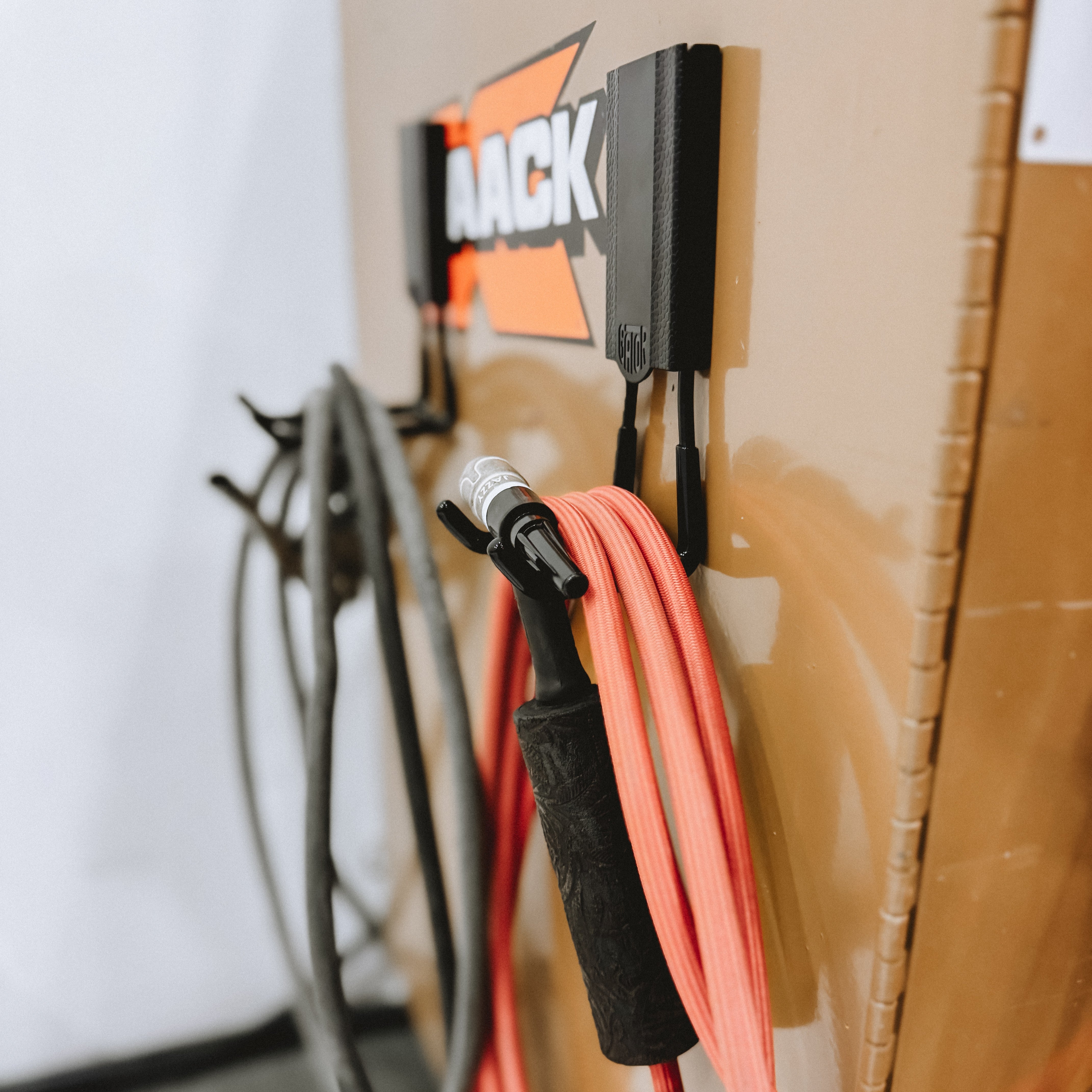 Two black magnetic hooks hold coiled red and gray cables by the side of a steel wall panel with an orange and white "AACK" logo.