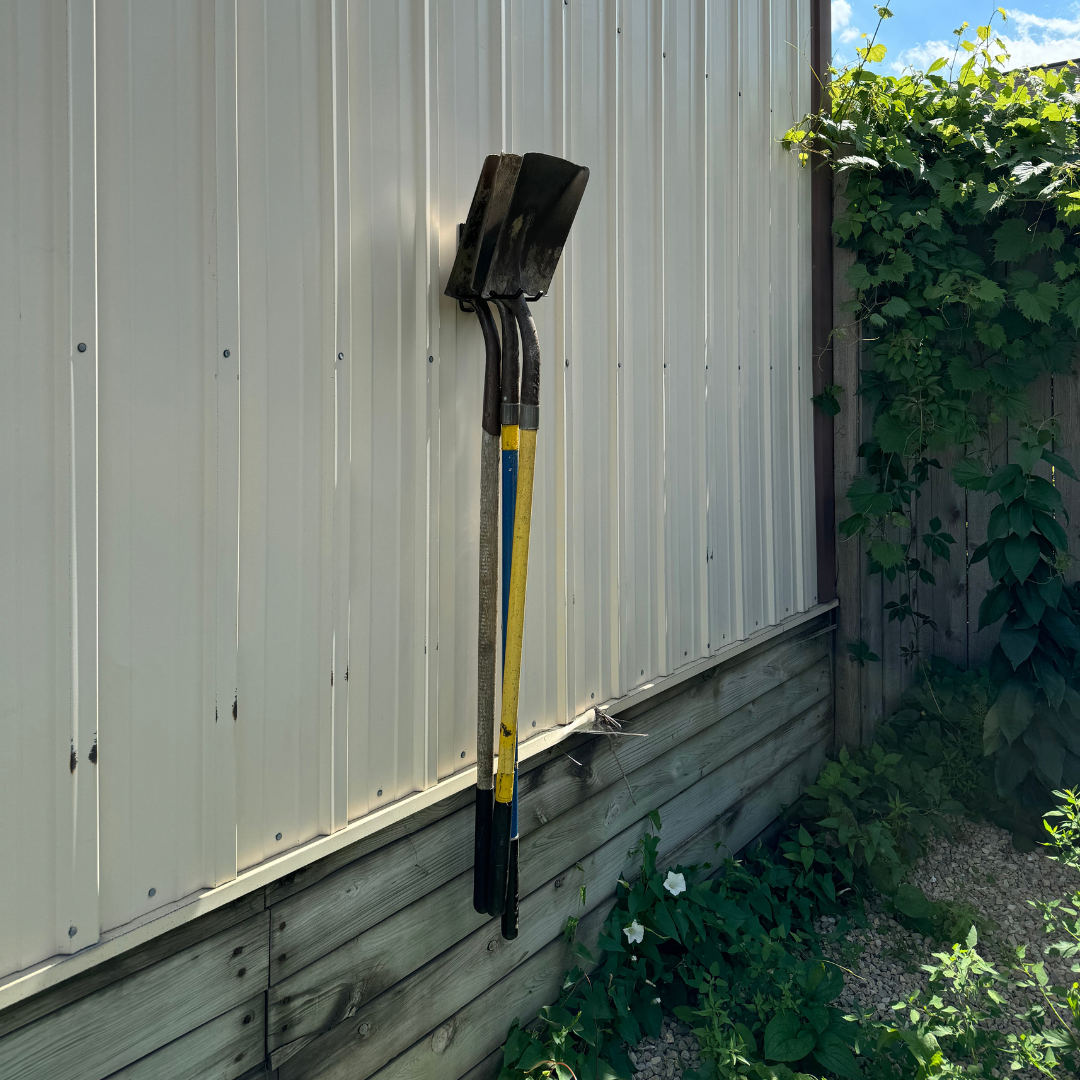 Two shovels with yellow and blue handles are hanging by red hooks on a vertical steel wall panel.