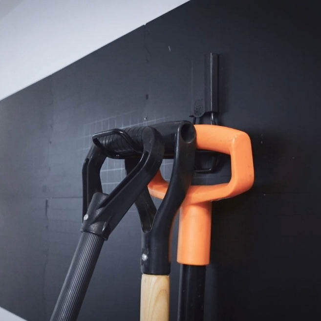 Two snow shovels with black and orange plastic handles are stored on red heavy-duty storage hooks attached to a flat black steel wall panel.