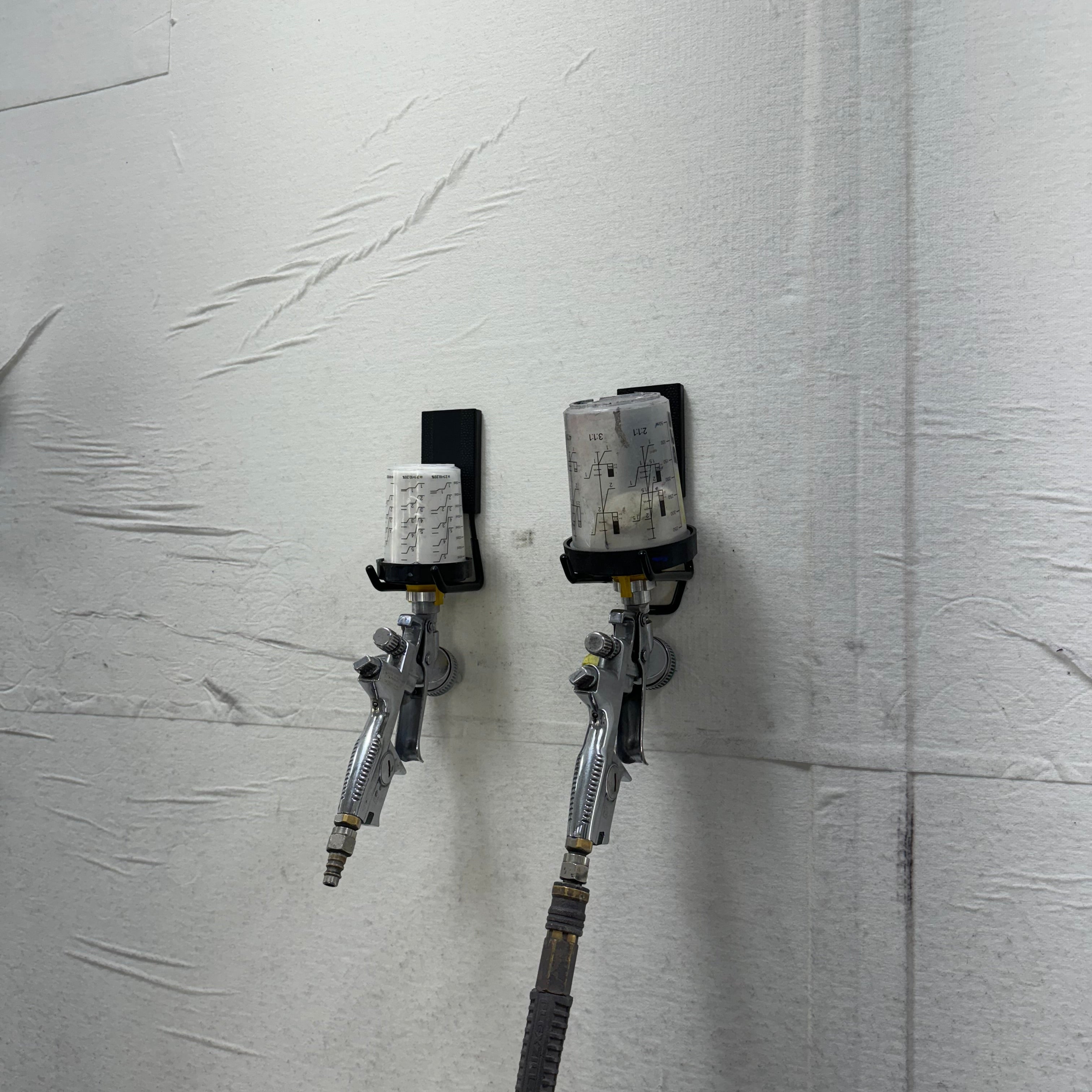Two spray guns with paint cups inverted and held by black magnetic hooks on a steel wall panel in a workshop.