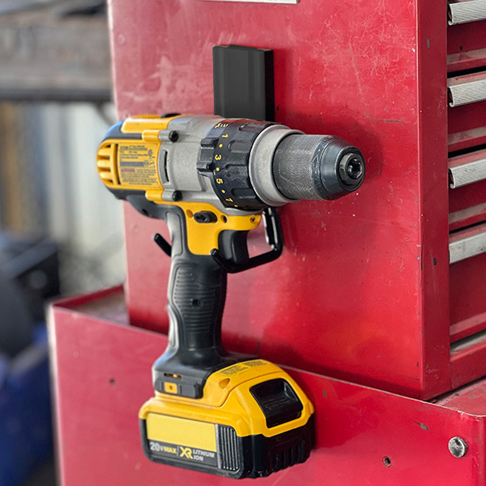 A yellow and black cordless drill is held by a black magnetic hook attached to a red steel toolbox in a workshop.