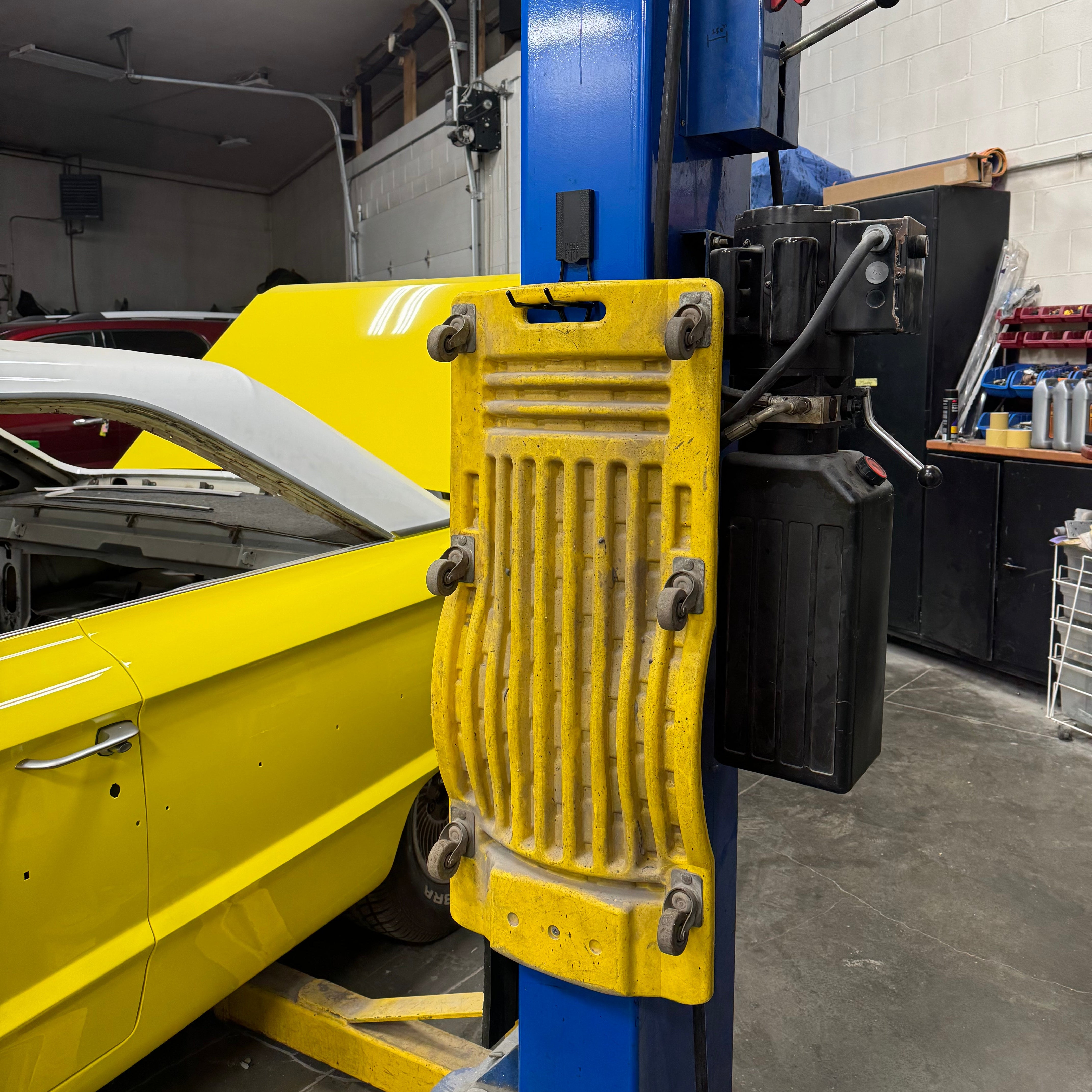 A yellow mechanic's creeper is hung on a blue steel post using a red Gator Magnetics magnetic hook in a garage with a yellow car nearby.