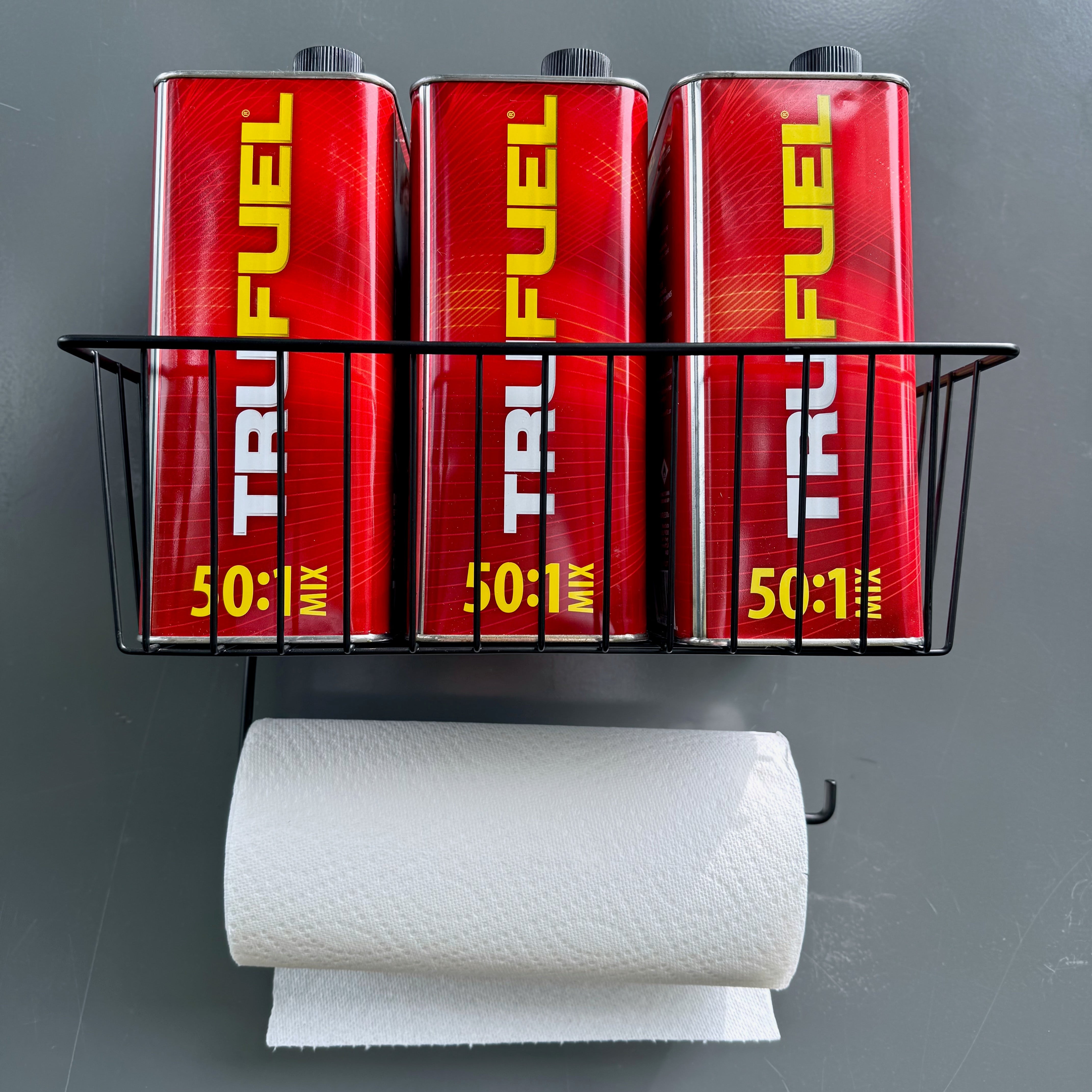 Magnetic basket for garage or workshop storage and organization, holding three TruFuel 50:1 mix fuel cans with a paper towel roll mounted below, keeping essential supplies easily accessible on a steel surface.